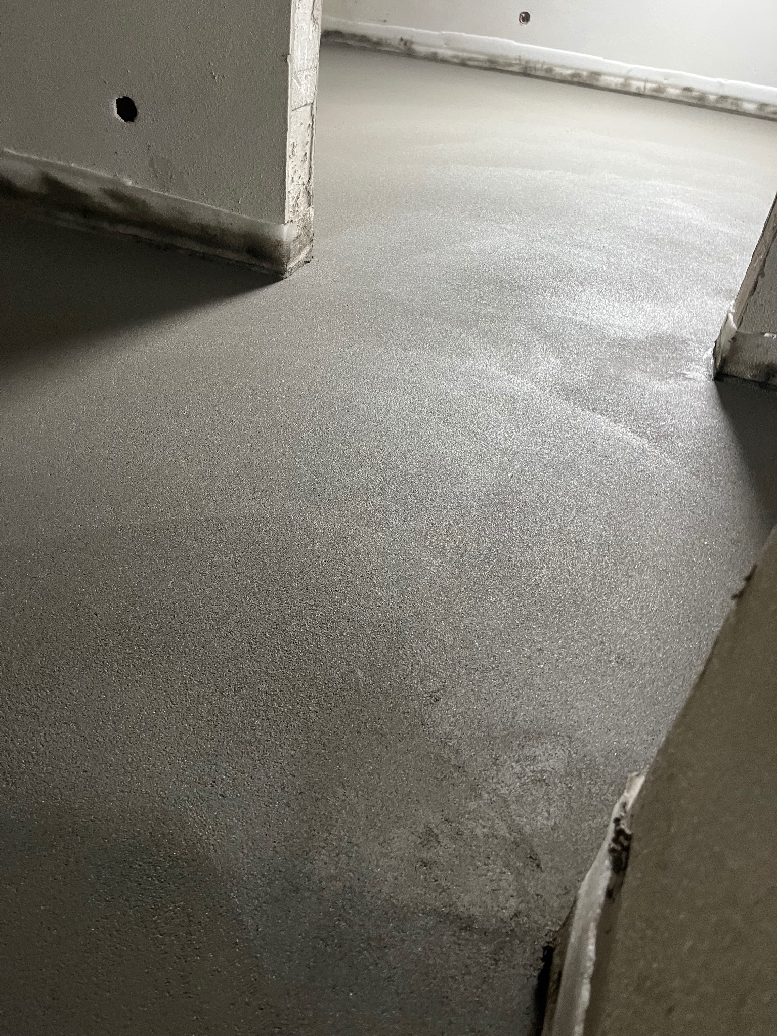 Świeżo wylana, szara posadzka w pomieszczeniu, widoczne fragmenty ścian i progu drzwi. Gładka powierzchnia, gotowa do dalszych prac wykończeniowych.
