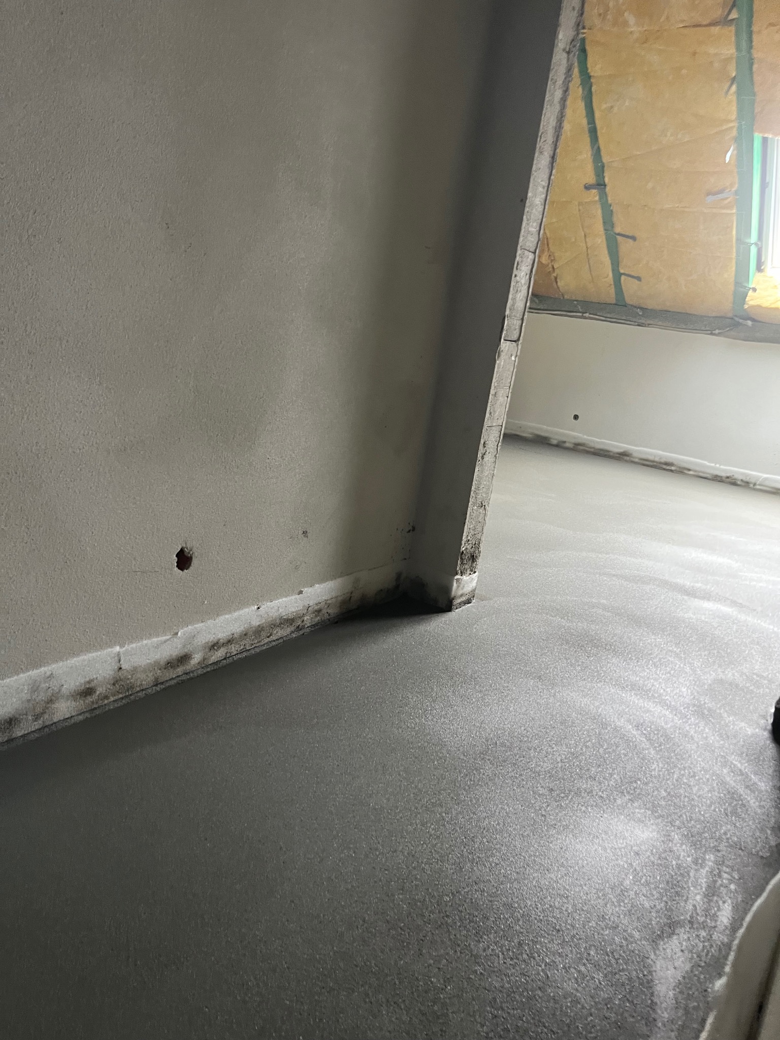 Świeżo wylana, szara posadzka w pomieszczeniu z widoczną izolacją poddasza i fragmentem okna. Ślady wilgoci na ścianie przy podłodze.
