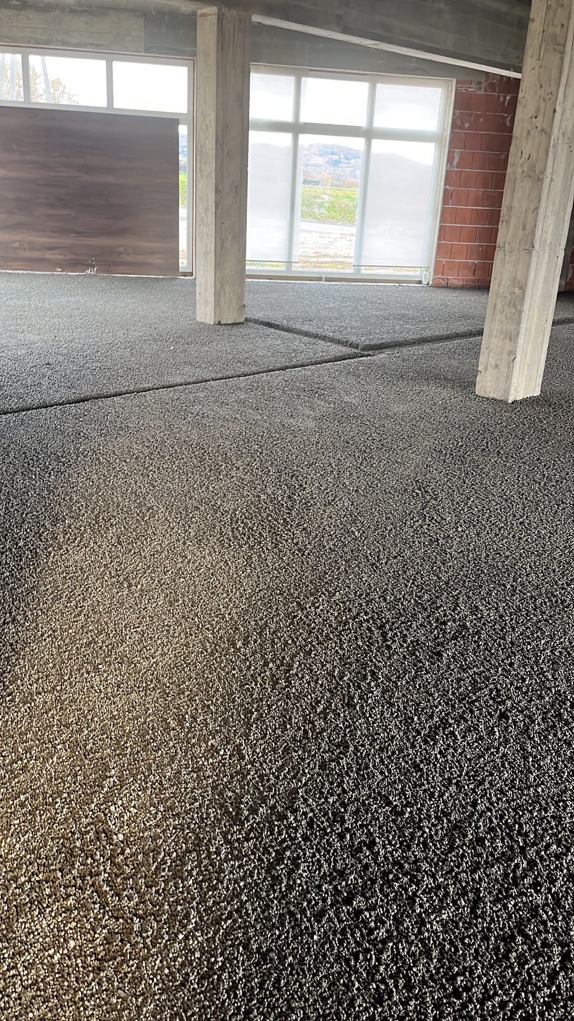 Szara, wylana posadzka w surowym wnętrzu z betonowymi kolumnami i dużymi oknami. Widoczne dylatacje. W tle widok na zewnątrz.