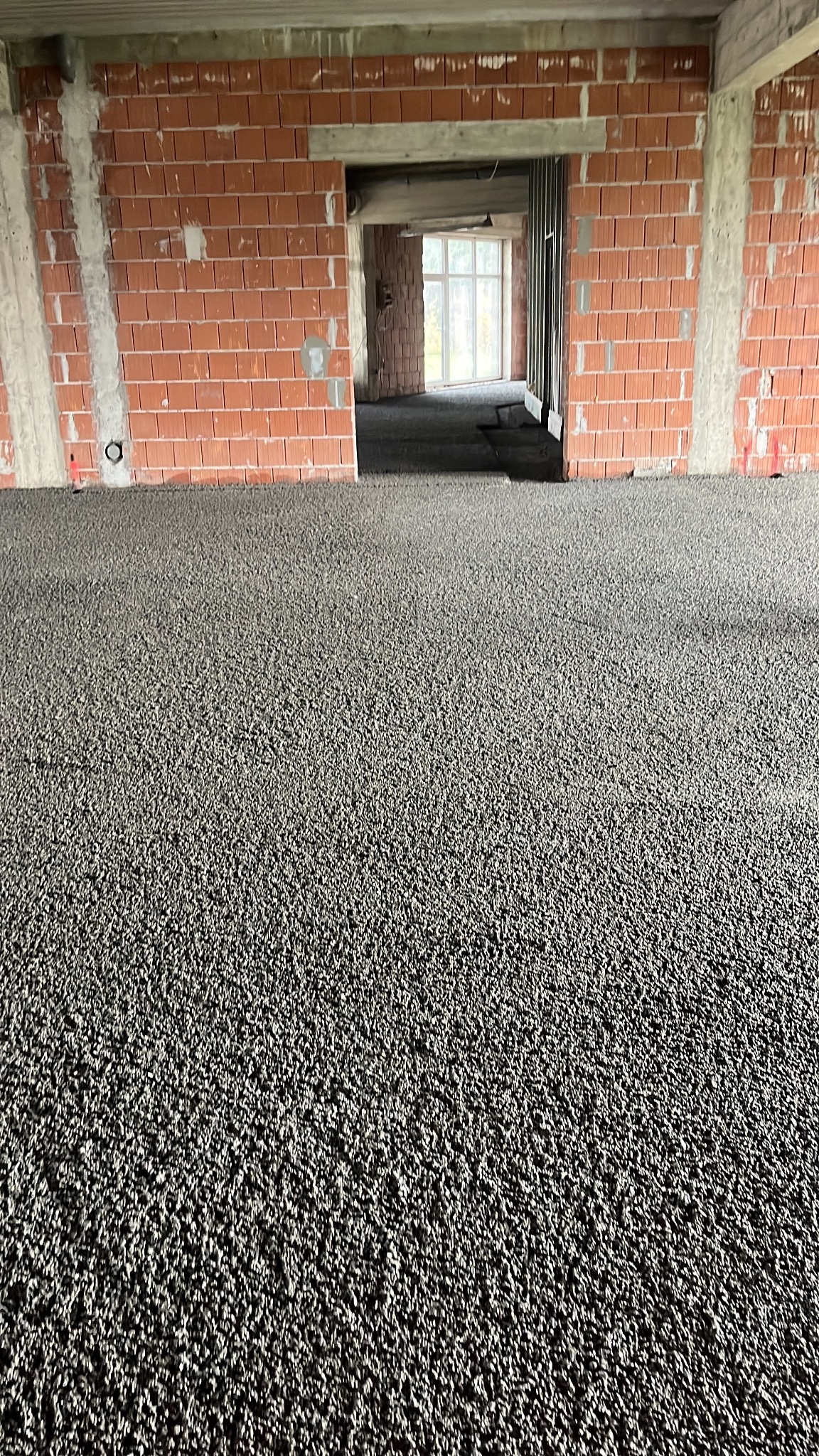 Wnętrze w trakcie budowy z wylaną, szarą posadzką. Widoczne ceglane ściany i otwór drzwiowy prowadzący do dalszej części pomieszczenia z oknem.
