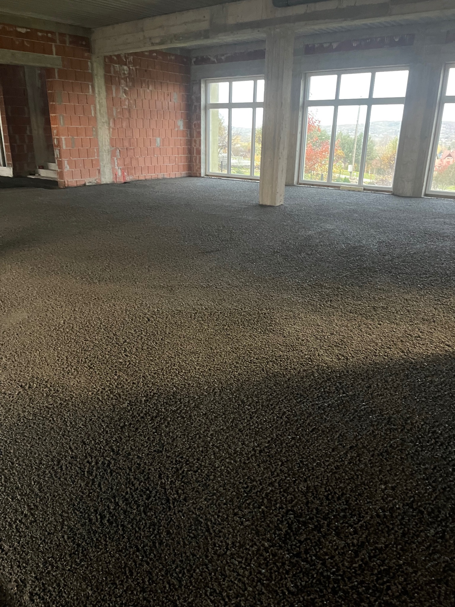 Wnętrze w stanie surowym z wylaną, ciemną posadzką. Widoczne okna z widokiem na zewnątrz i ceglane ściany. Betonowe słupy podtrzymują strop.