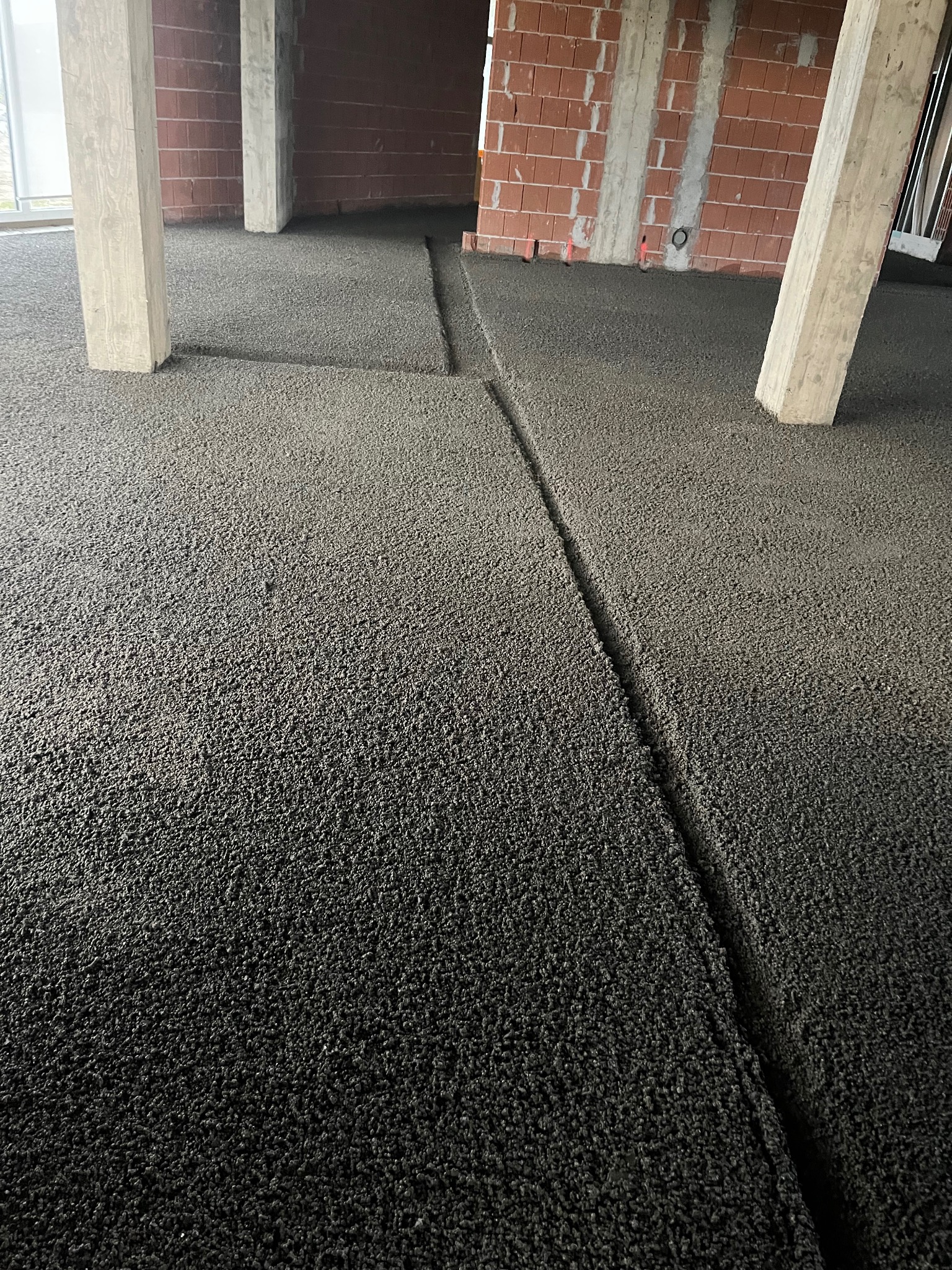 Wylana, ciemna posadzka w surowym wnętrzu budynku w trakcie budowy. Widoczne betonowe kolumny i ściany z czerwonej cegły. Szczelina dylatacyjna w posadzce.