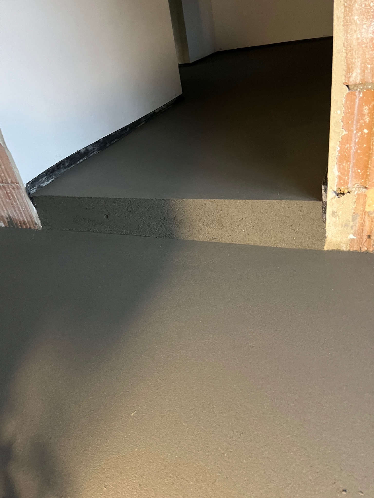 Świeżo wylana, gładka, szara posadzka w pomieszczeniu z fragmentami ceglanych ścian i białej ściany. Widoczny etap prac wykończeniowych.