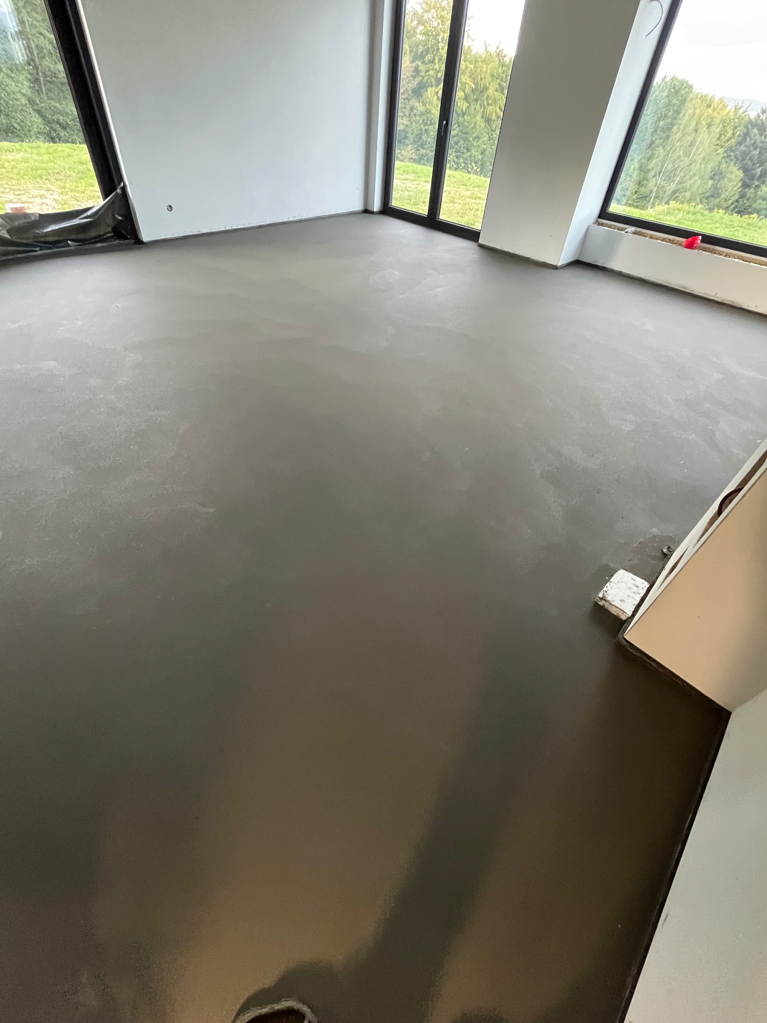 Gładka, szara posadzka w jasnym wnętrzu z dużymi oknami. Widoczne zabezpieczenia okien folią. Minimalistyczny design, świeżo wylana powierzchnia.