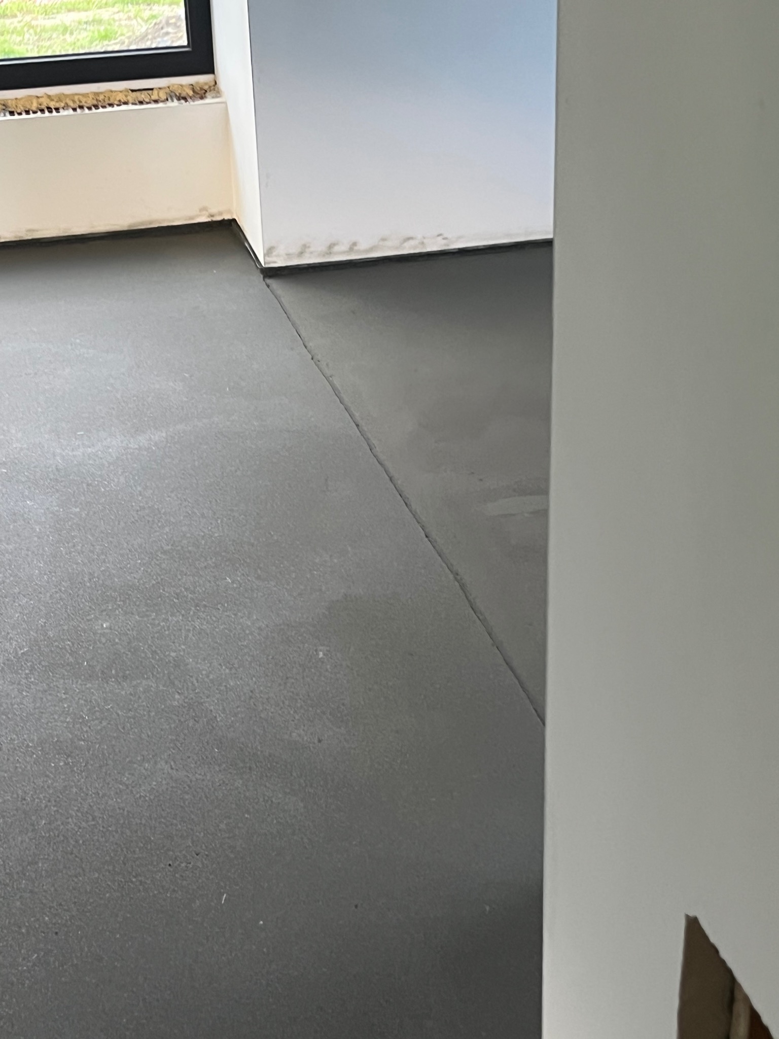 Szara, gładka posadzka w jasnym wnętrzu, widoczna przy oknie i ścianie. Minimalistyczny design, świeżo wylana, z delikatną teksturą i widocznymi łączeniami.