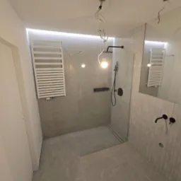 Nowoczesna łazienka z kabiną prysznicową typu walk-in, grzejnikiem drabinkowym i czarną armaturą. Minimalistyczny design z szarymi płytkami i podświetleniem LED.