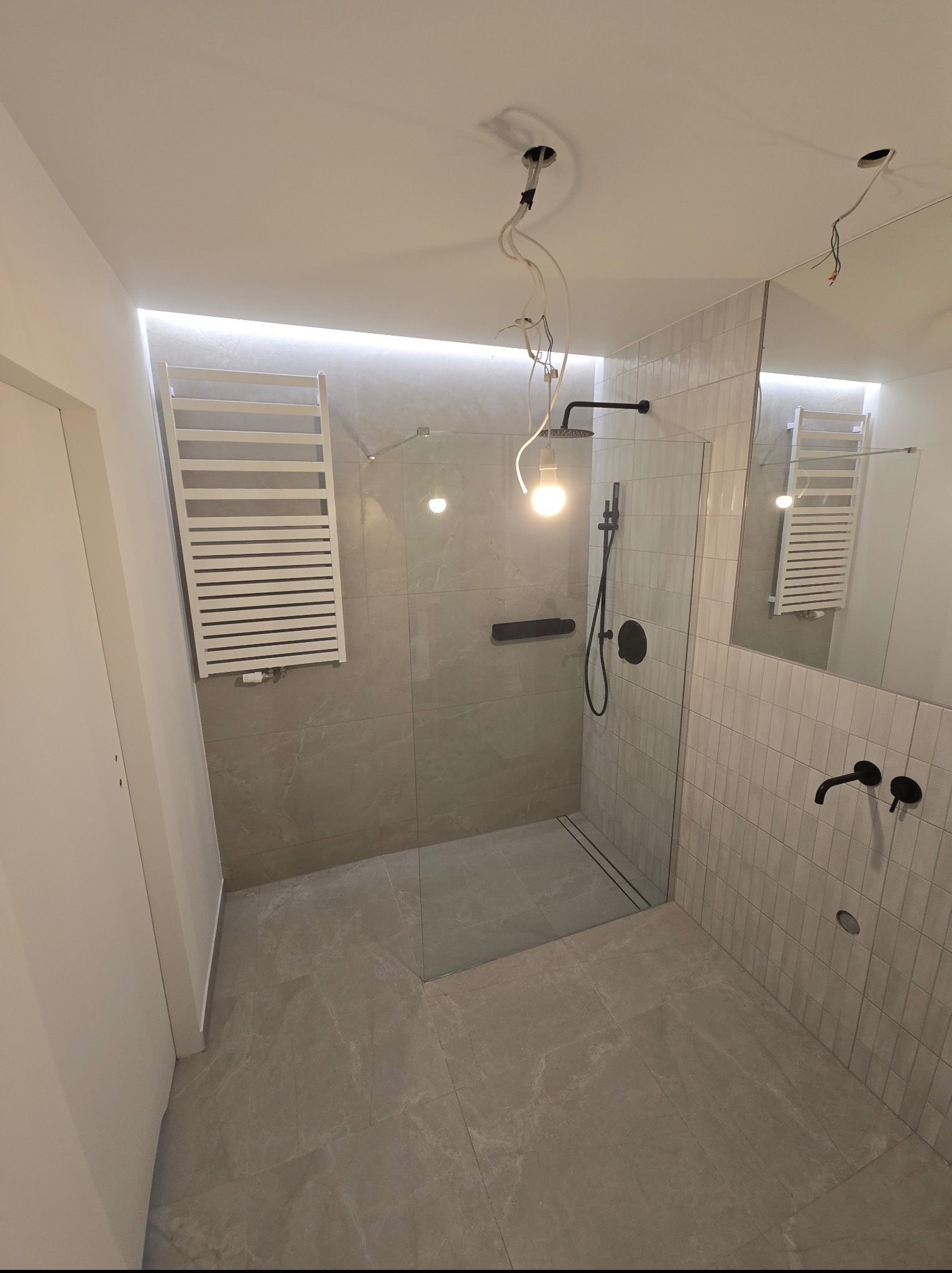 Nowoczesna łazienka z kabiną prysznicową typu walk-in, grzejnikiem drabinkowym i czarną armaturą. Minimalistyczny design z szarymi płytkami i podświetleniem LED.