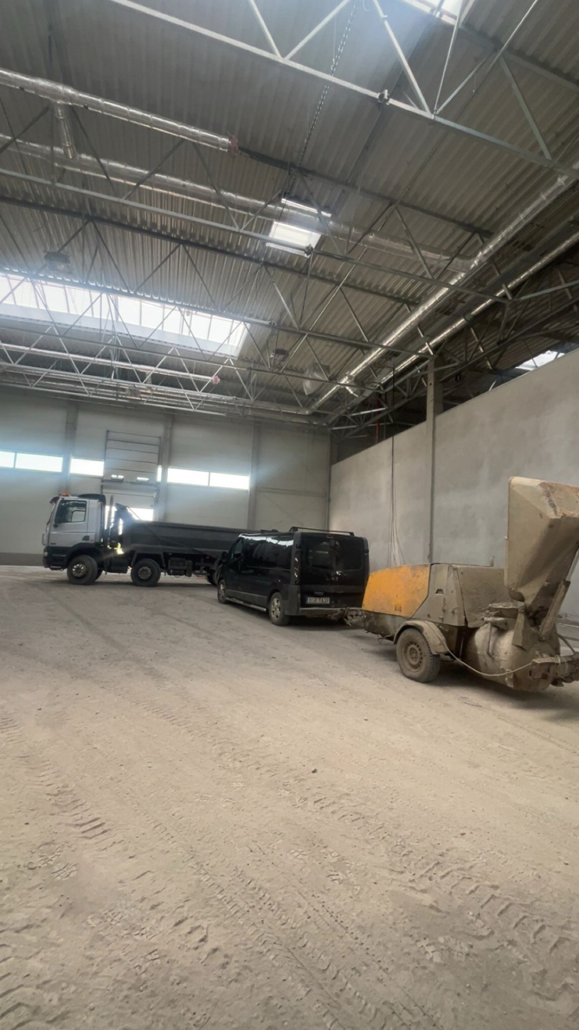 Wnętrze hali z pojazdami: ciężarówka, bus i maszyna do wylewek. Widoczne ślady opon na podłożu. Oświetlenie górne, surowy beton na ścianach.