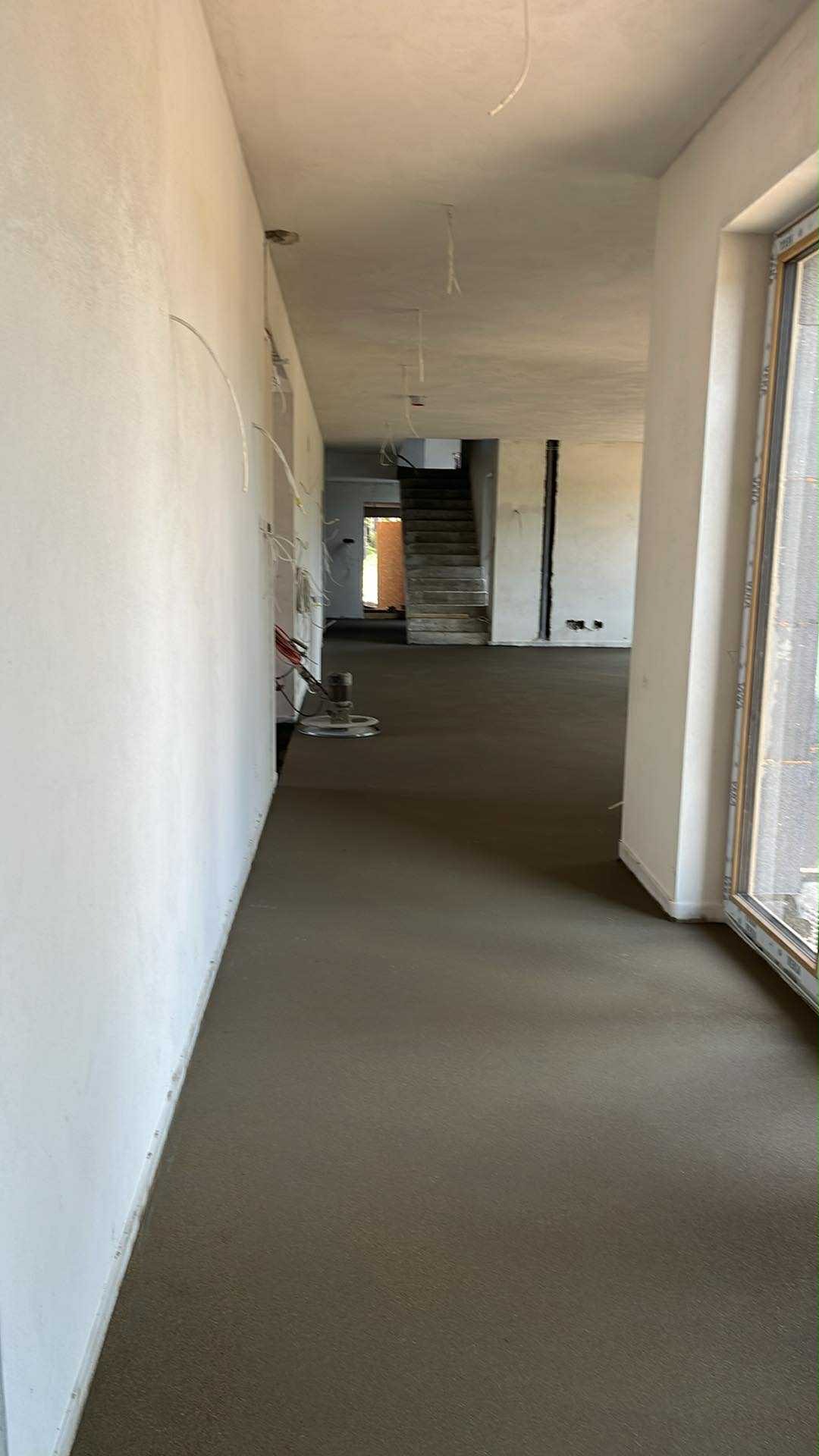 Świeża, szara wylewka podłogowa w surowym wnętrzu domu jednorodzinnego, widok na schody i okno. W tle maszyna do wygładzania powierzchni.