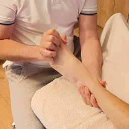 BLASK NATURY GRZEGORZ JASIAK - Masażysta w białej koszulce masuje stopę pacjenta na leżance z ręcznikiem, widoczne dłonie masujące i fragment drewnianej podłogi.