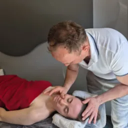 Mężczyzna wykonuje masaż twarzy kobiety leżącej na stole z czerwonym ręcznikiem, w gabinecie z szarym tłem. Dłonie masażysty delikatnie dotykają skóry.