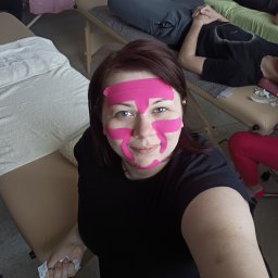 BLASK NATURY GRZEGORZ JASIAK - Kobieta z plastrami kinesiotaping na twarzy, uśmiechnięta, na tle gabinetu z leżankami i pacjentami. Widoczne taśmy w kolorze różowym.