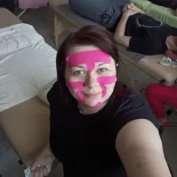 Kobieta z plastrami kinesiotaping na twarzy, uśmiechnięta, na tle gabinetu z leżankami i pacjentami. Widoczne taśmy w kolorze różowym.