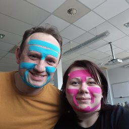 BLASK NATURY GRZEGORZ JASIAK - Uśmiechnięta para z kolorowymi taśmami kinesiotaping na twarzach, w jasnym wnętrzu z widocznym projektorem i oświetleniem biurowym.