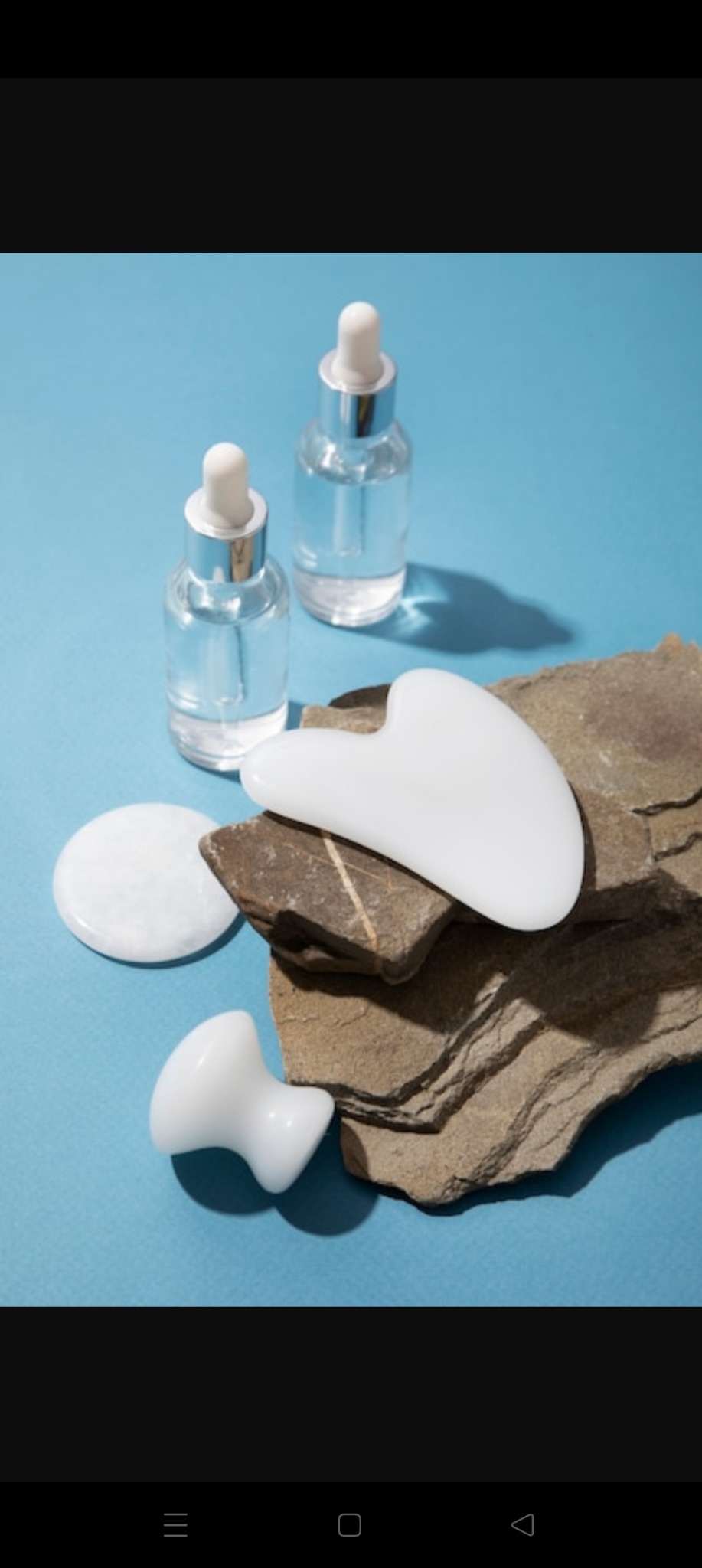 Zestaw do pielęgnacji: serum w szklanych butelkach z pipetą, białe kamienie gua sha i roller na tle błękitu i naturalnego kamienia.
