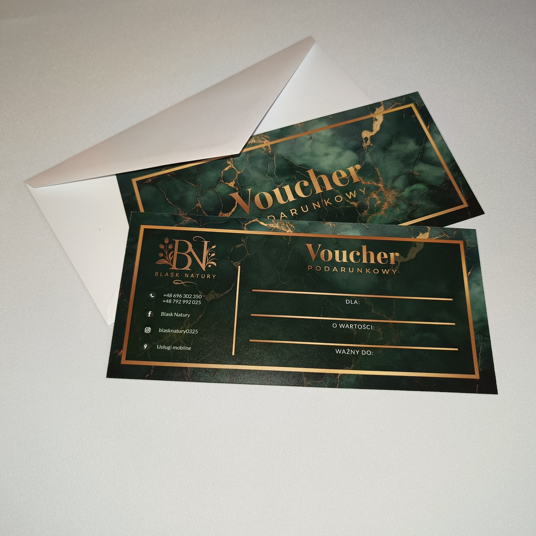 Elegancki voucher podarunkowy Blask Natury w odcieniach zieleni i złota, leżący obok białej koperty. Idealny prezent na zabiegi kosmetyczne i relaksacyjne.