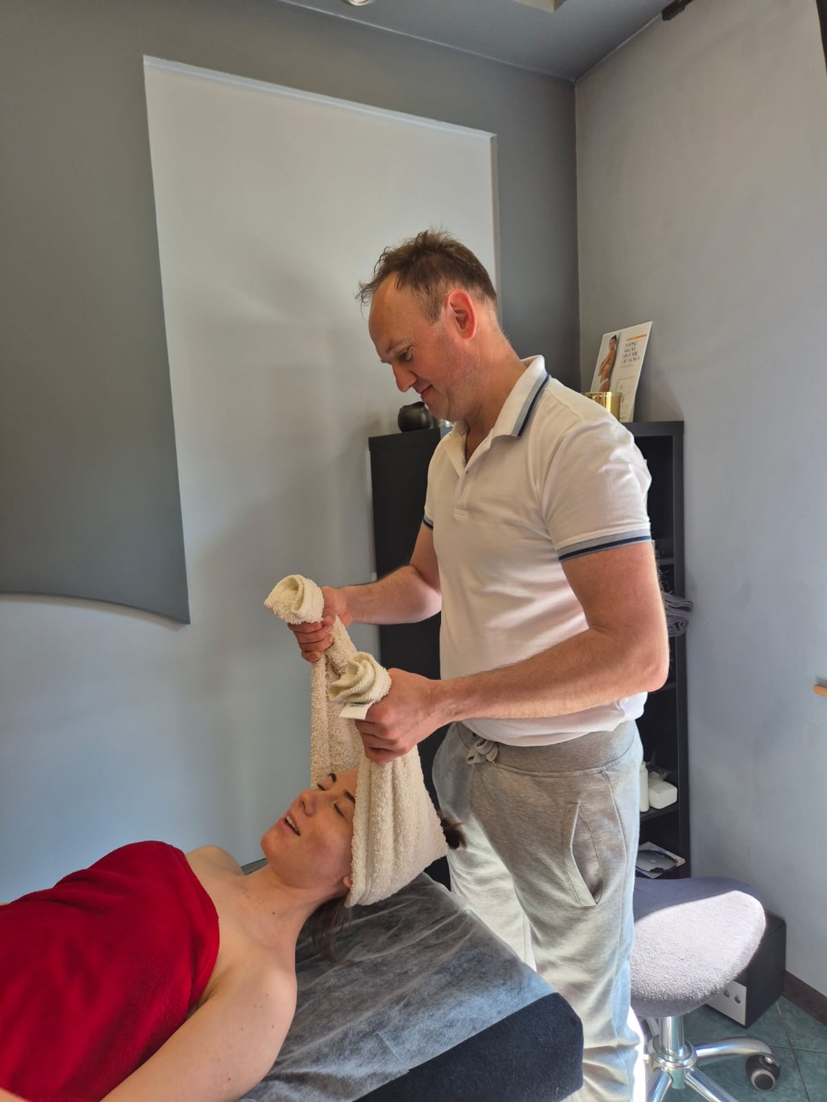 Mężczyzna w białej koszulce wykonuje masaż głowy ciepłym ręcznikiem kobiecie leżącej na stole do masażu w gabinecie. Czerwony ręcznik okrywa ciało.