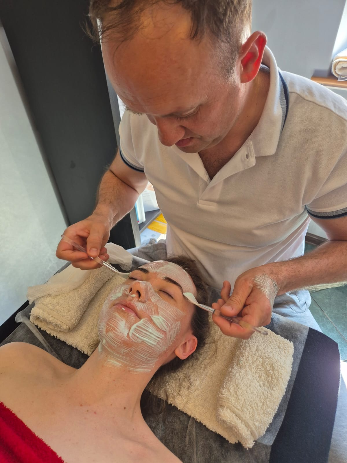 Nakładanie maseczki na twarz kobiety za pomocą pędzli. Kosmetyczka w białej koszulce pochyla się nad klientką leżącą na leżance w salonie kosmetycznym.