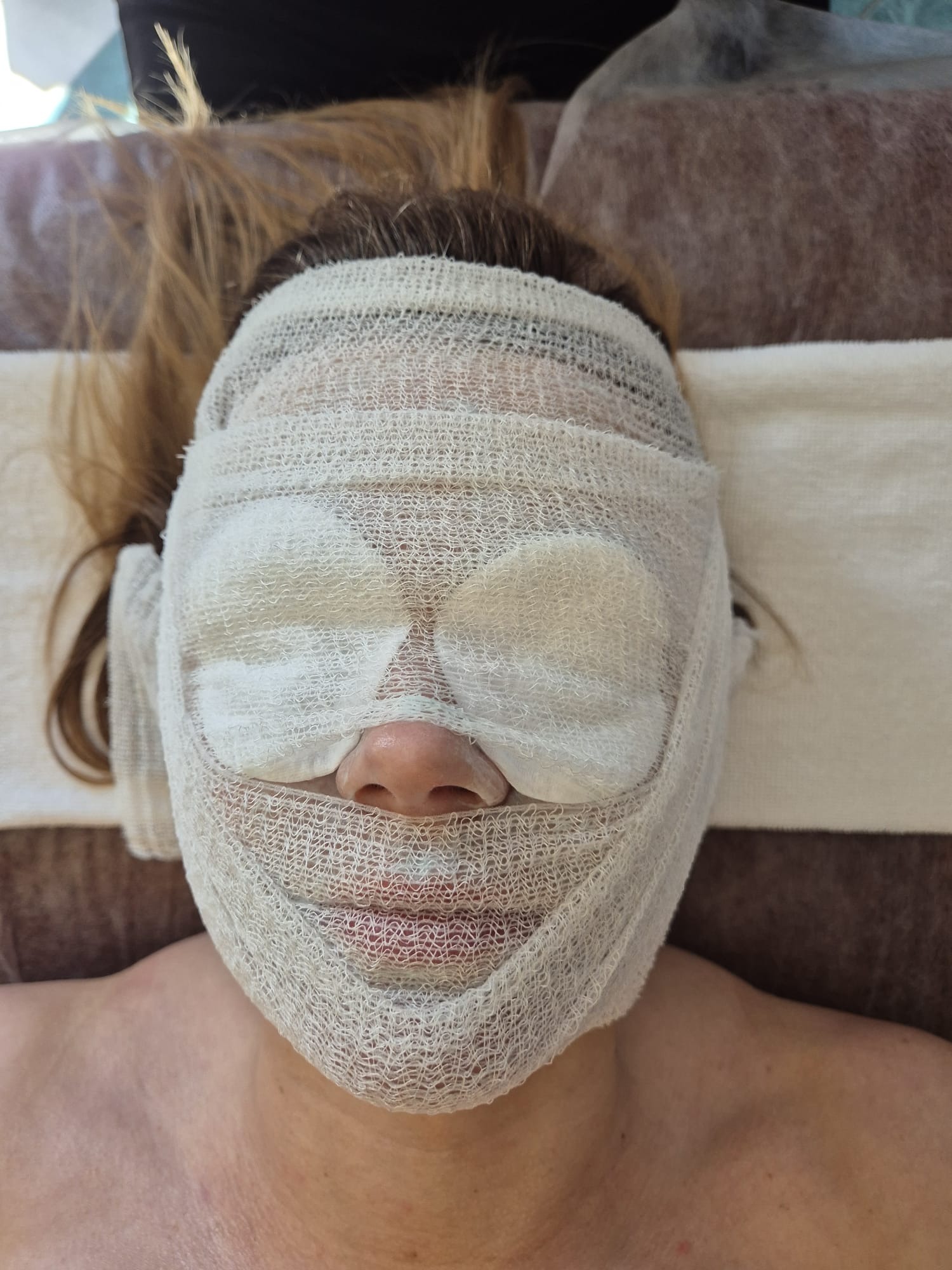 Kobieta leży na plecach z twarzą owiniętą w gazę i waciki na oczach, przygotowana do zabiegu kosmetycznego. Widok z góry, neutralne oświetlenie.