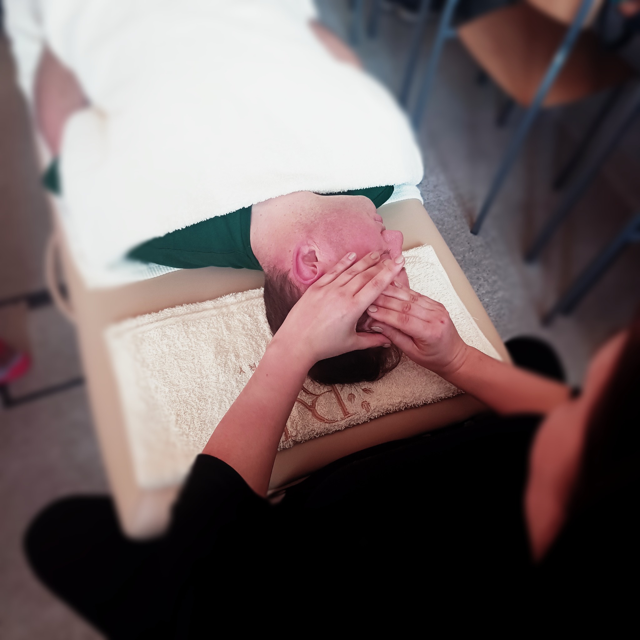 Masaż twarzy wykonywany na leżącym kliencie, z użyciem rąk masażysty. Ujęcie z góry, widoczny ręcznik i fragment kozetki do masażu.