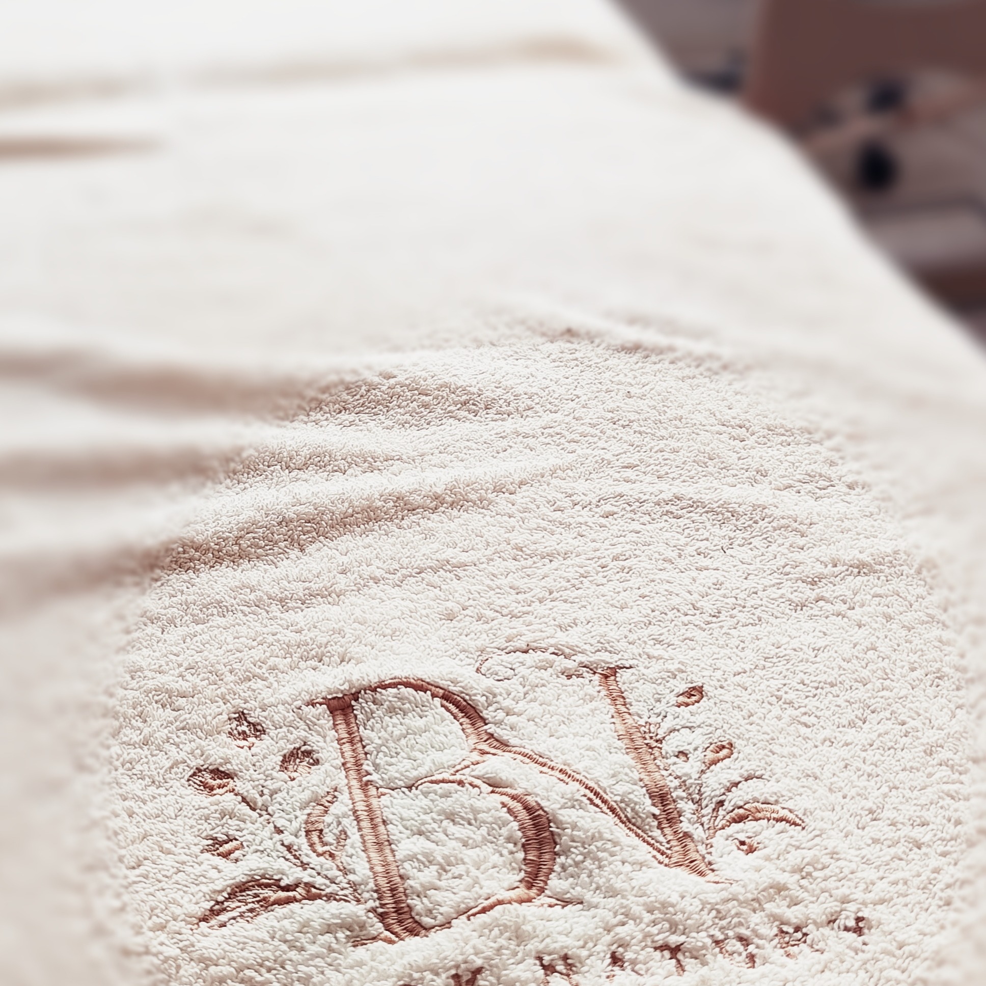 Jasny, puszysty ręcznik z wyhaftowanymi inicjałami 'BV' i delikatnymi motywami roślinnymi, leżący na stole w salonie kosmetycznym.