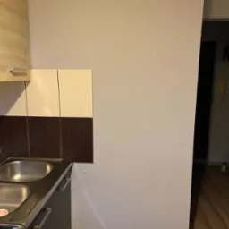 Kuchnia po remoncie: szare ściany, zabudowa z brązowo-beżowymi płytkami nad zlewem, fragment podłogi z ciemnego drewna. Widok na fragment korytarza.