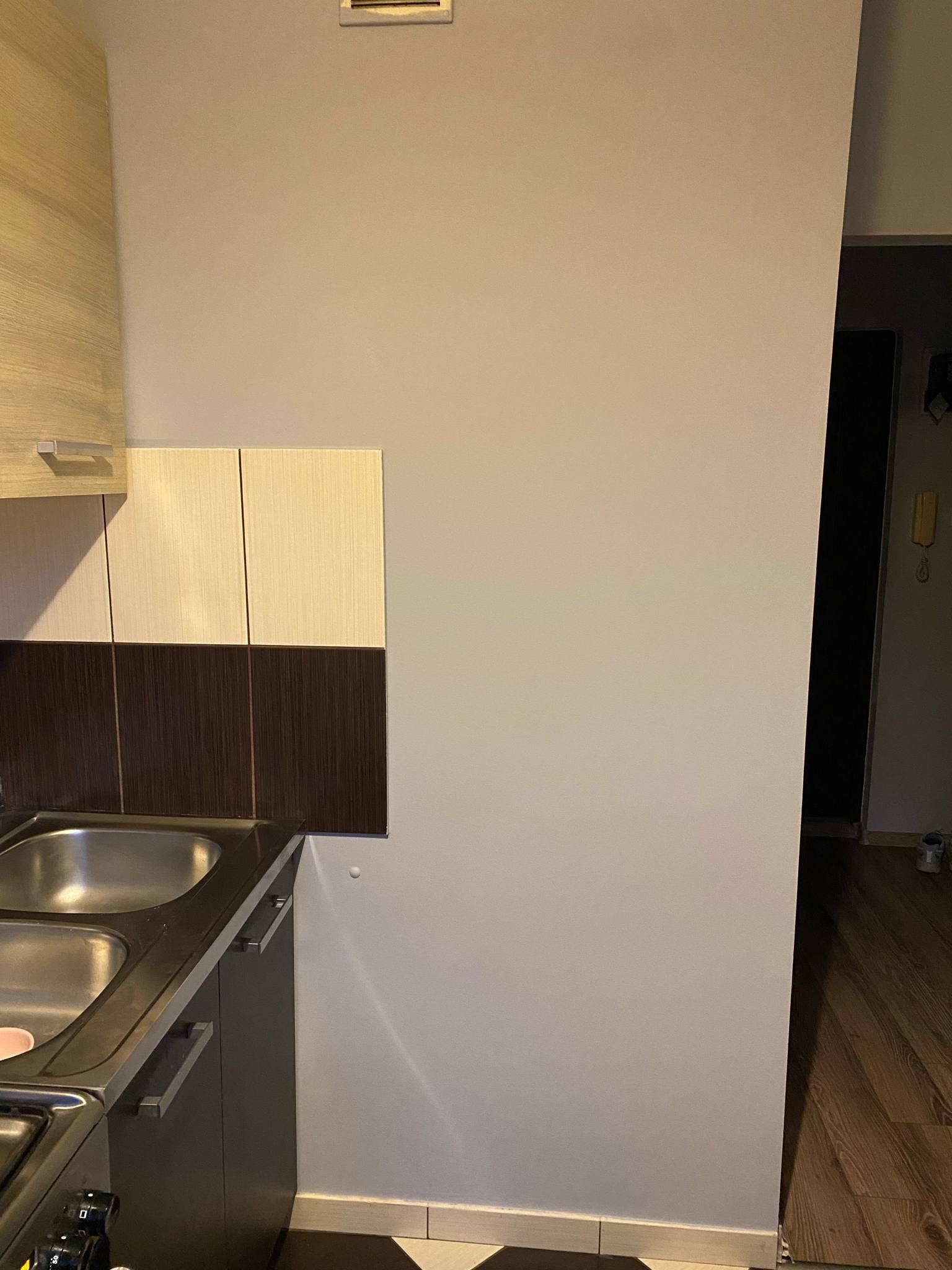 Kuchnia po remoncie: szare ściany, zabudowa z brązowo-beżowymi płytkami nad zlewem, fragment podłogi z ciemnego drewna. Widok na fragment korytarza.