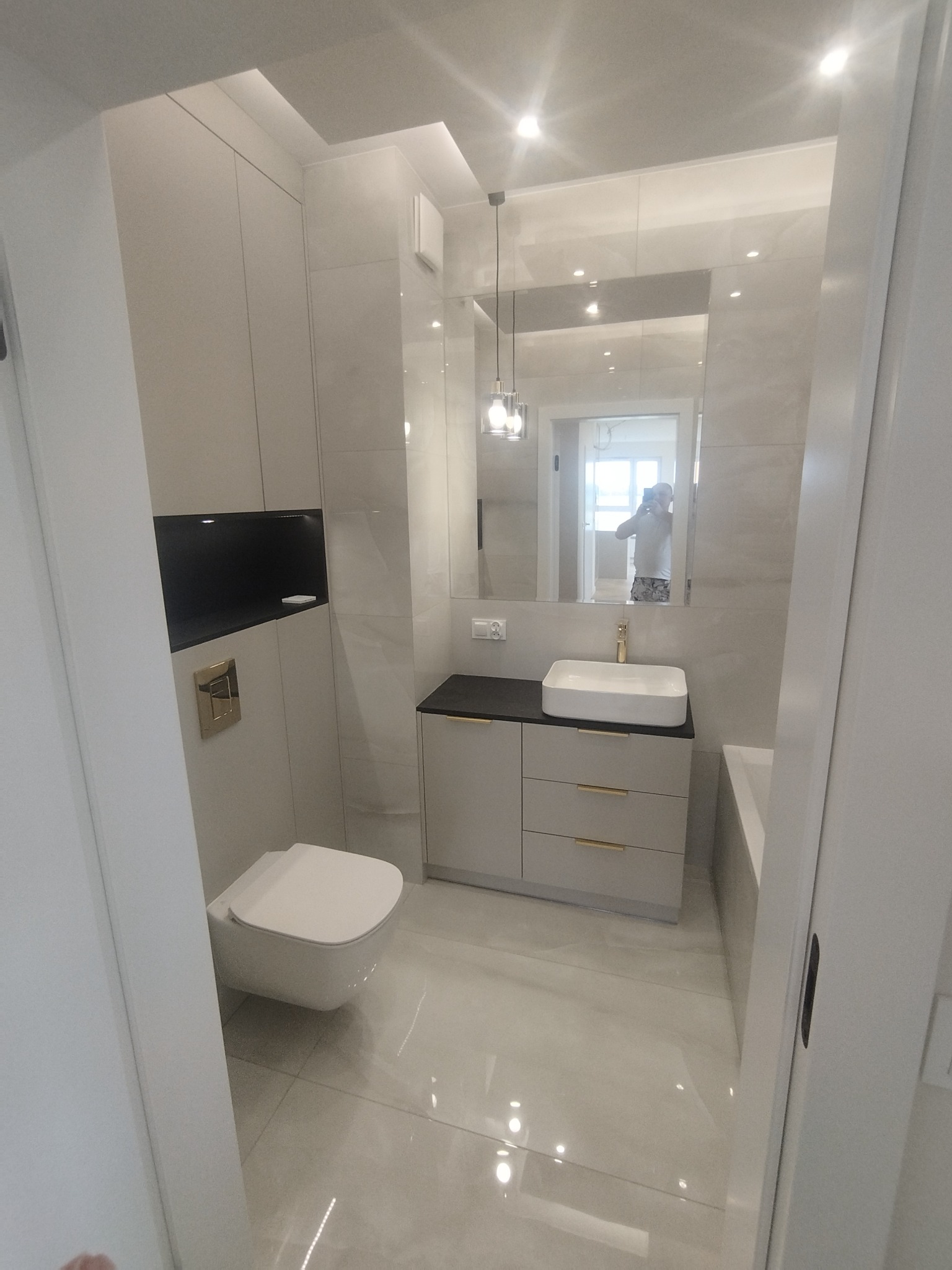 Nowoczesna łazienka w jasnych barwach z podwieszaną toaletą, szafką z umywalką i lustrem. Widoczne płytki imitujące marmur i minimalistyczne oświetlenie.