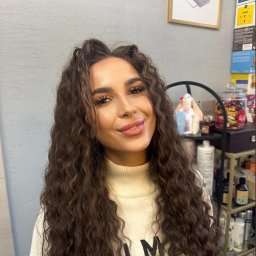 Alena Semiantsova - Salon fryzjerski, Fryzjer Warszawa - Portret kobiety z burzą brązowych, kręconych włosów po zabiegu stylizacji w salonie, w tle ściana i półka z kosmetykami. Uśmiech, makijaż.