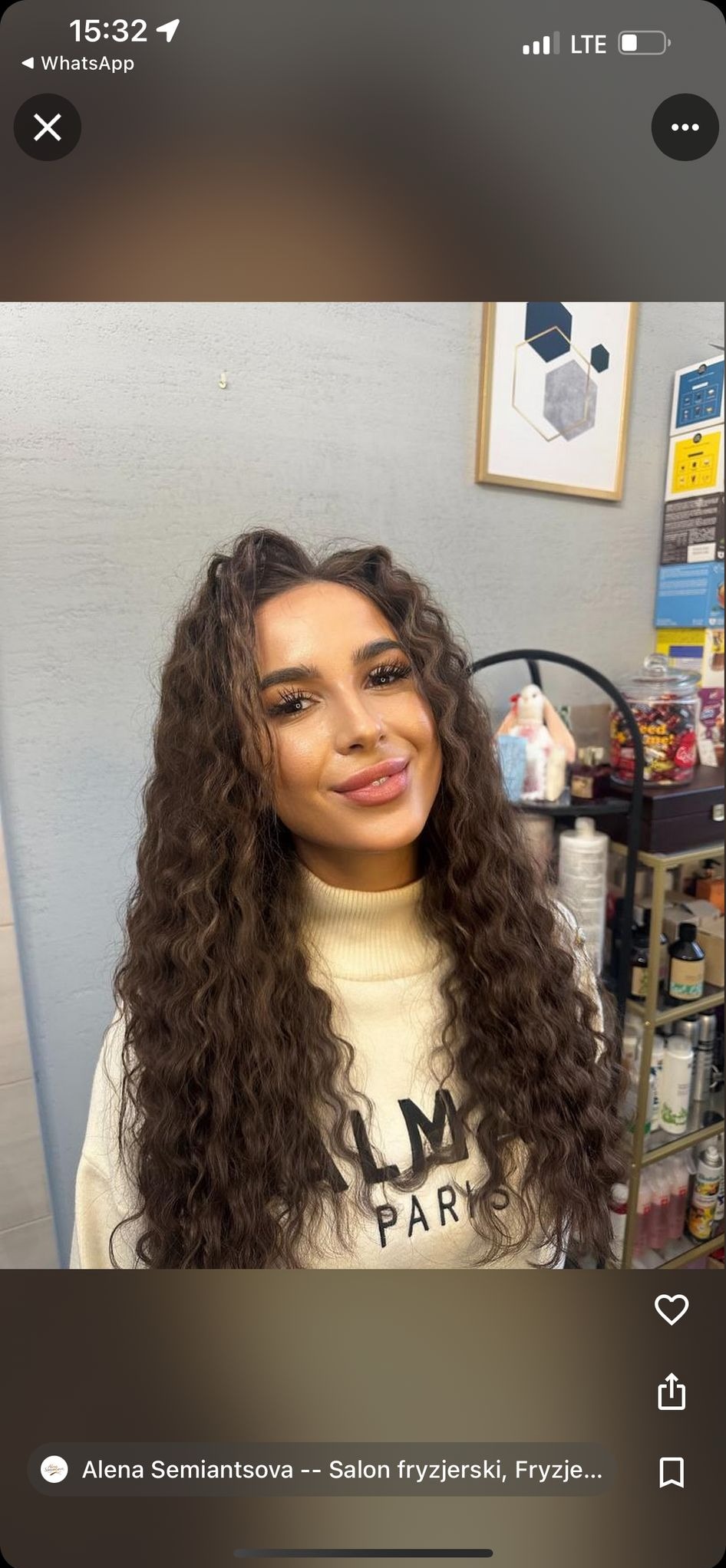 Portret kobiety z burzą brązowych, kręconych włosów po zabiegu stylizacji w salonie, w tle ściana i półka z kosmetykami. Uśmiech, makijaż.