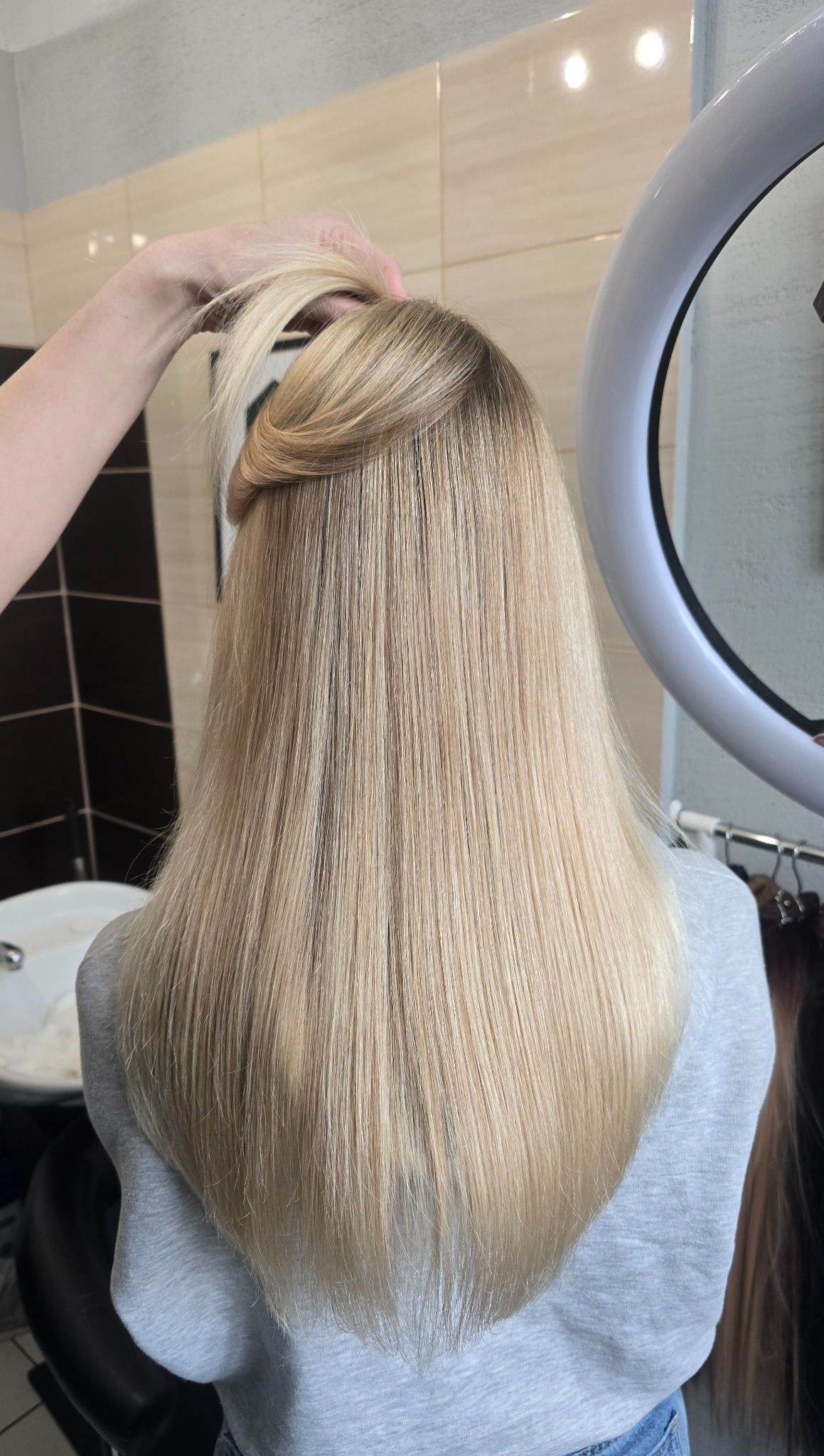 Blond włosy po koloryzacji AirTouch. Subtelne przejścia kolorów, naturalny efekt. W tle ring doświetlający i wieszak z doczepianymi pasmami włosów. Fryzjer.