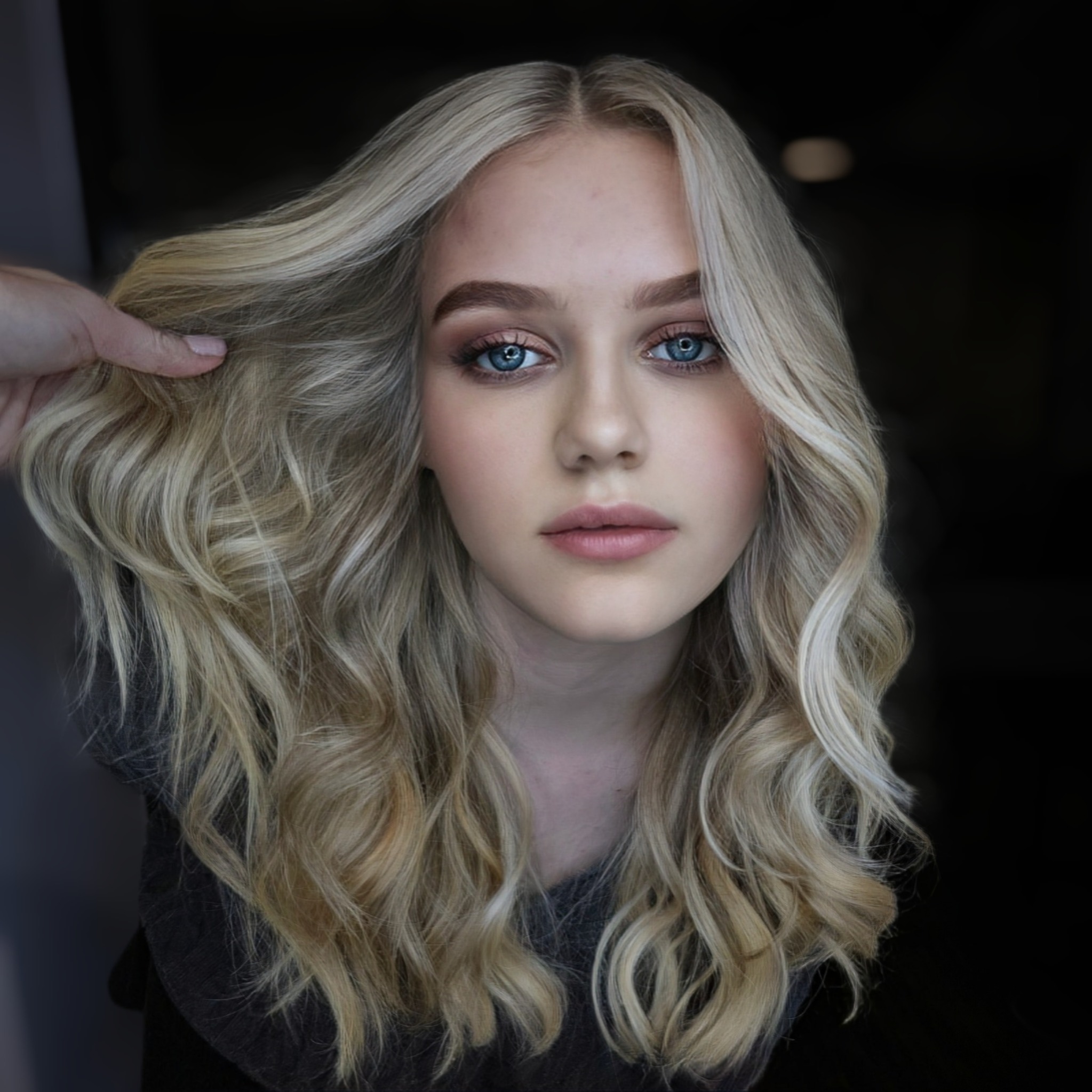 Portret młodej kobiety z blond włosami w stylu AirTouch, z delikatnym rozświetleniem i naturalnym blondem, wykonanym w salonie fryzjerskim. Widoczna dłoń stylisty.