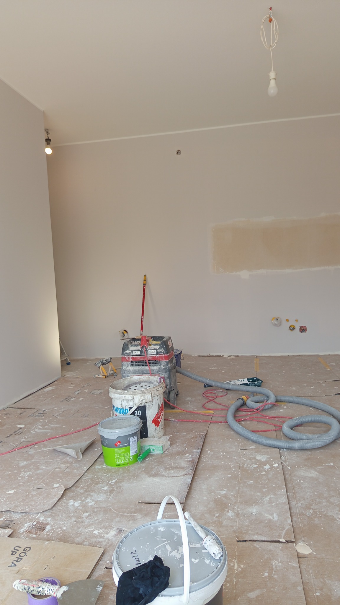 Przygotowane do malowania wnętrze z widocznym sprzętem, wiadrami farb i zabezpieczonym podłożem. Na ścianie widoczne ślady po szpachlowaniu.