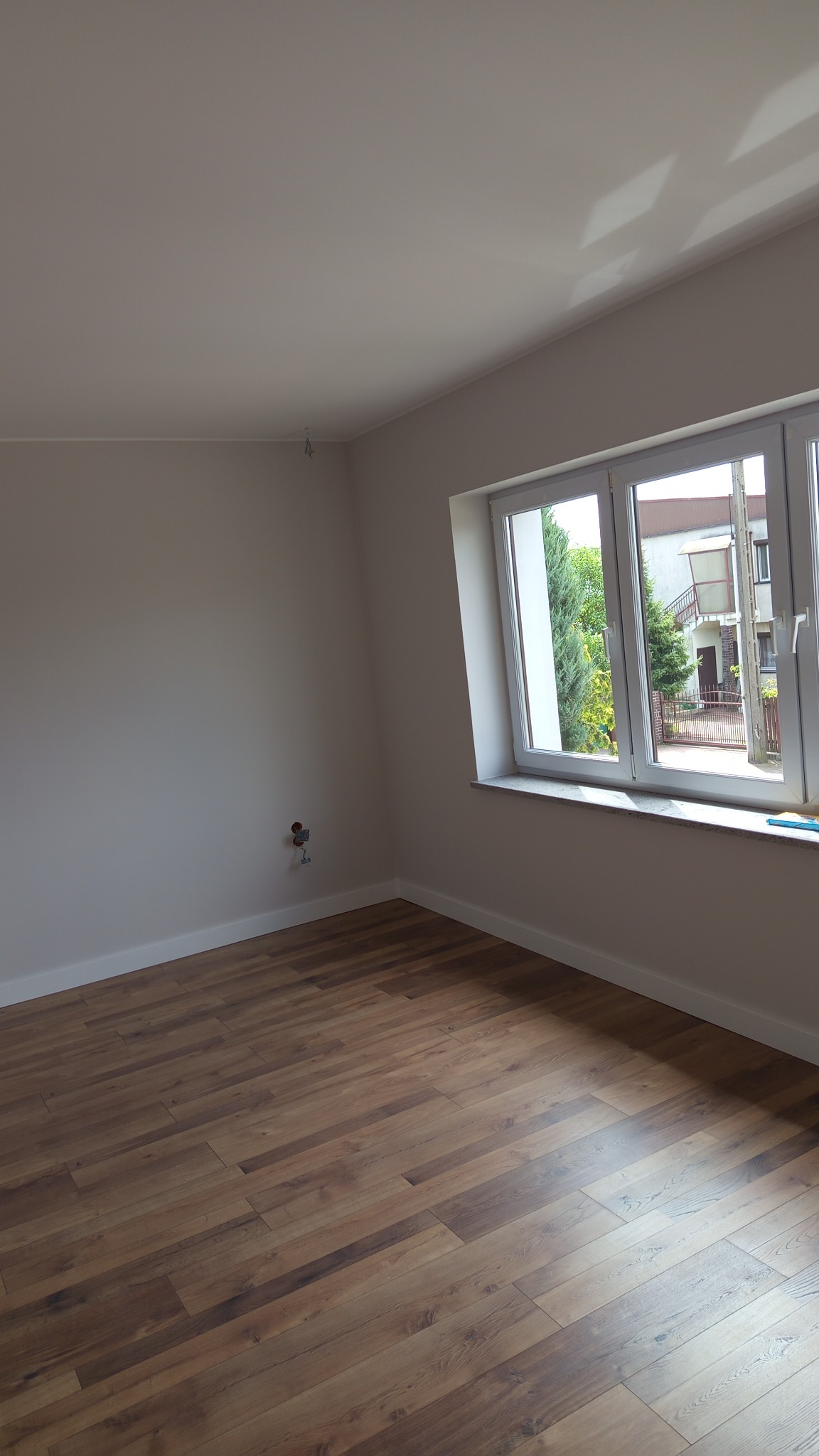 Jasne, puste wnętrze po remoncie: świeżo pomalowane ściany w odcieniach szarości, drewniana podłoga i białe listwy przypodłogowe. Widoczne okno z widokiem na zieleń.