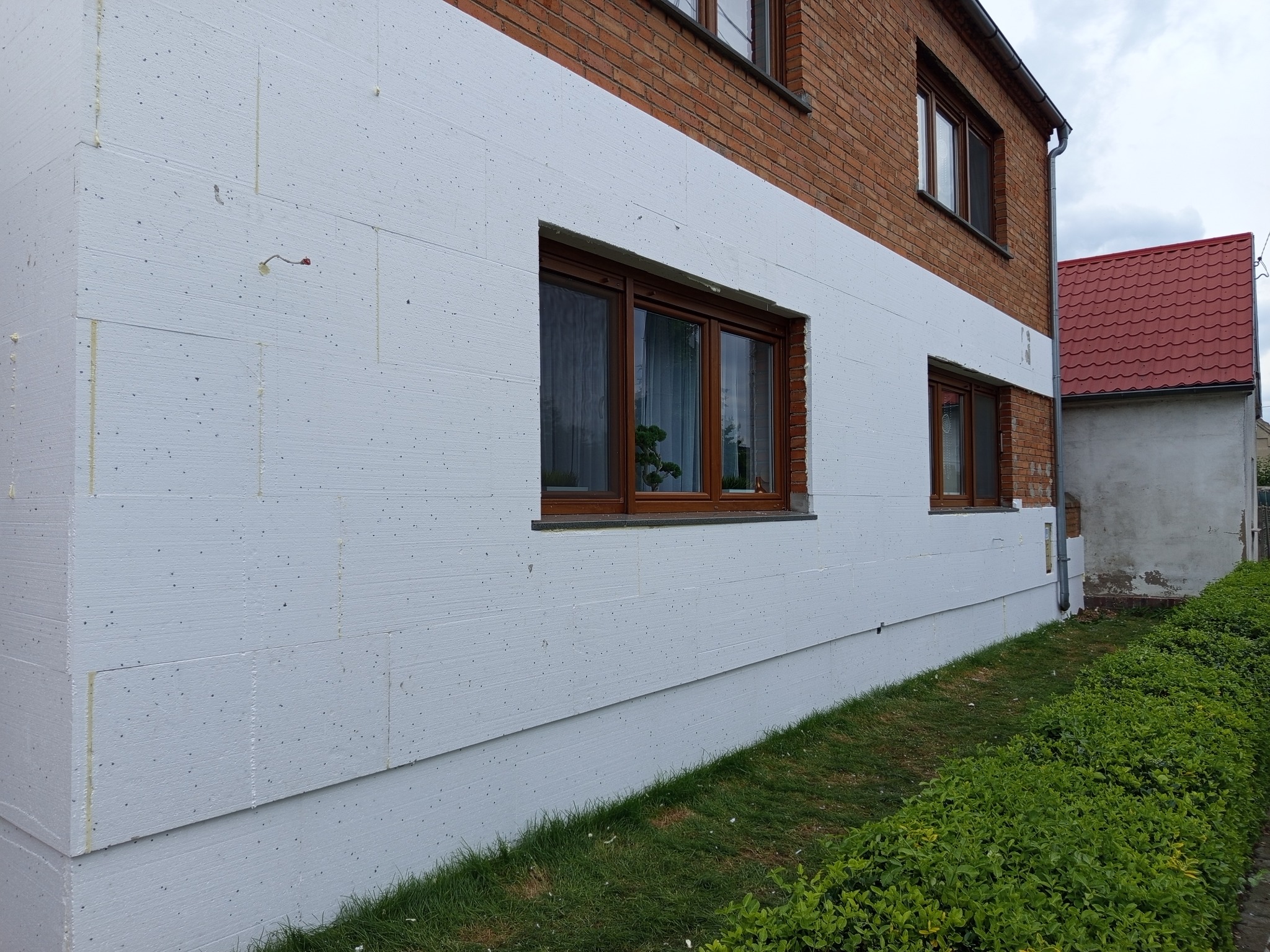 Ocieplenie budynku styropianem, widoczne płyty izolacyjne na elewacji, brązowe ramy okienne. Częściowo ocieplony budynek z czerwonej cegły.