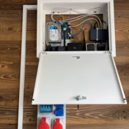 Otwarta, biała szafka z dyskiem twardym, routerem i okablowaniem na ścianie z drewnianych paneli, obok gniazda elektryczne. Elementy systemu monitoringu.