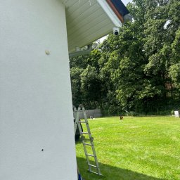 H&A SAFE SERVICE SYLWIA NITECKA - Kamera monitoringu zamontowana pod okapem domu z ogrodem w tle. Aluminiowa drabina oparta o ścianę. Widok na trawnik i zieleń. Dzień.