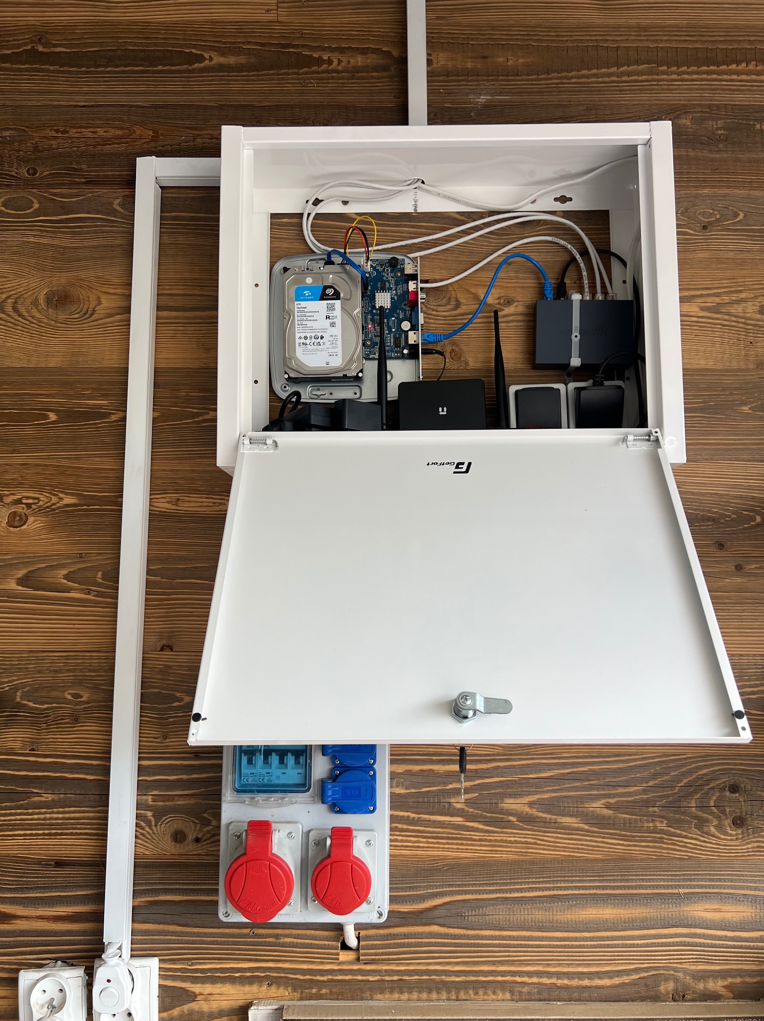 Otwarta, biała szafka z dyskiem twardym, routerem i okablowaniem na ścianie z drewnianych paneli, obok gniazda elektryczne. Elementy systemu monitoringu.