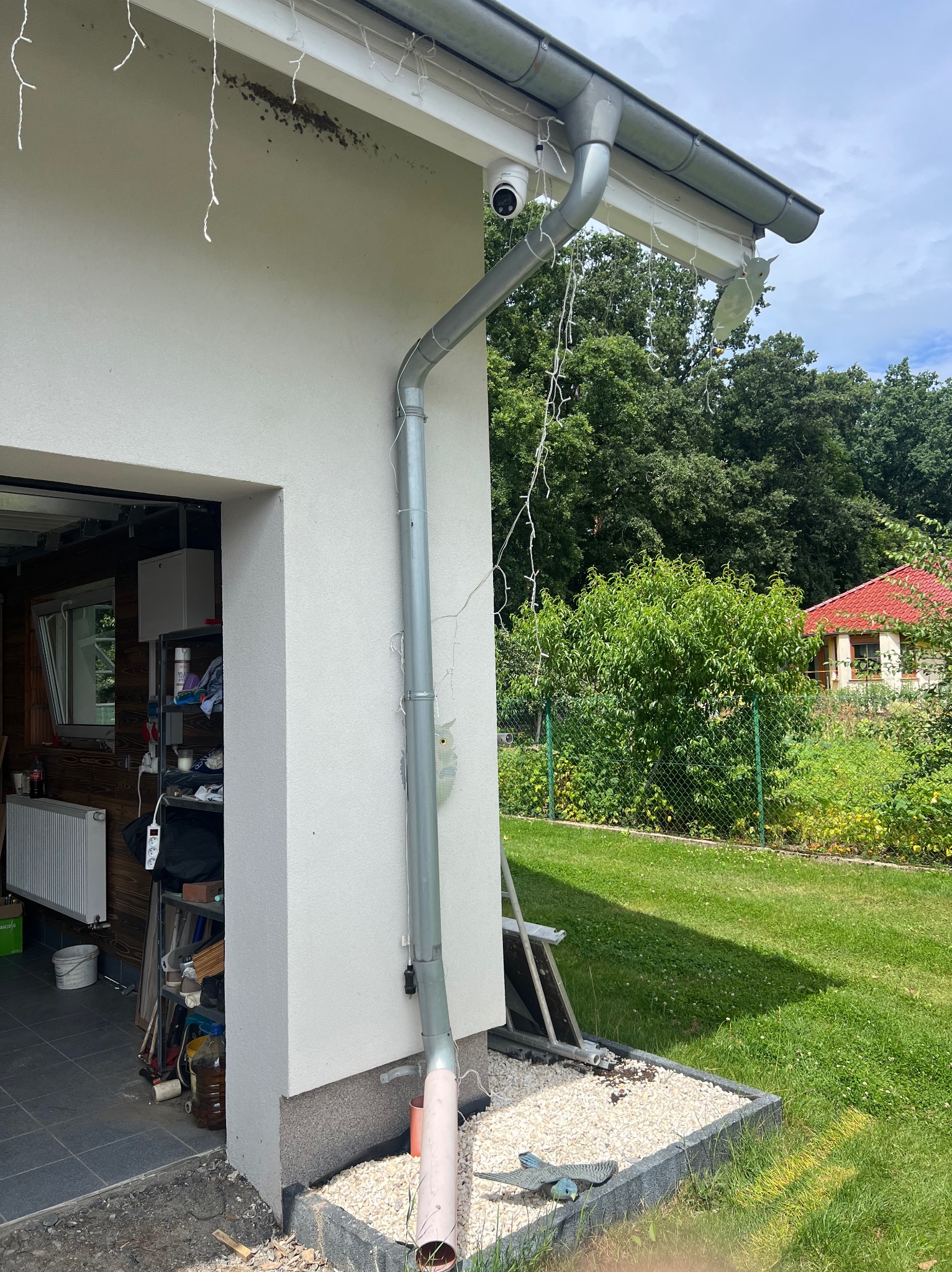 Biała kamera monitoringu zamontowana pod rynną domu z dekoracyjnymi lampkami, z widocznym odpływem rynnowym i fragmentem ogrodu w tle.