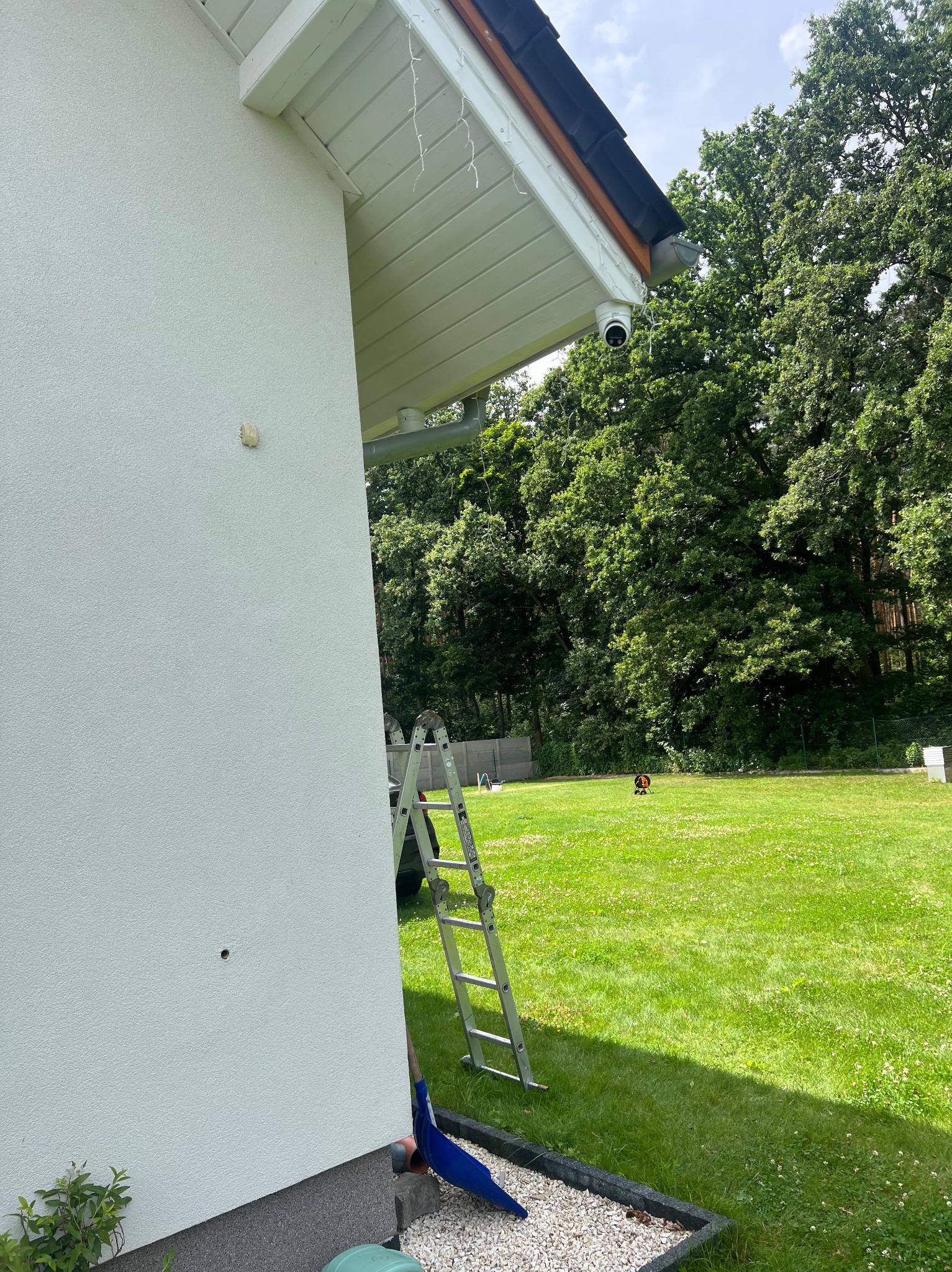 Kamera monitoringu zamontowana pod okapem domu z ogrodem w tle. Aluminiowa drabina oparta o ścianę. Widok na trawnik i zieleń. Dzień.