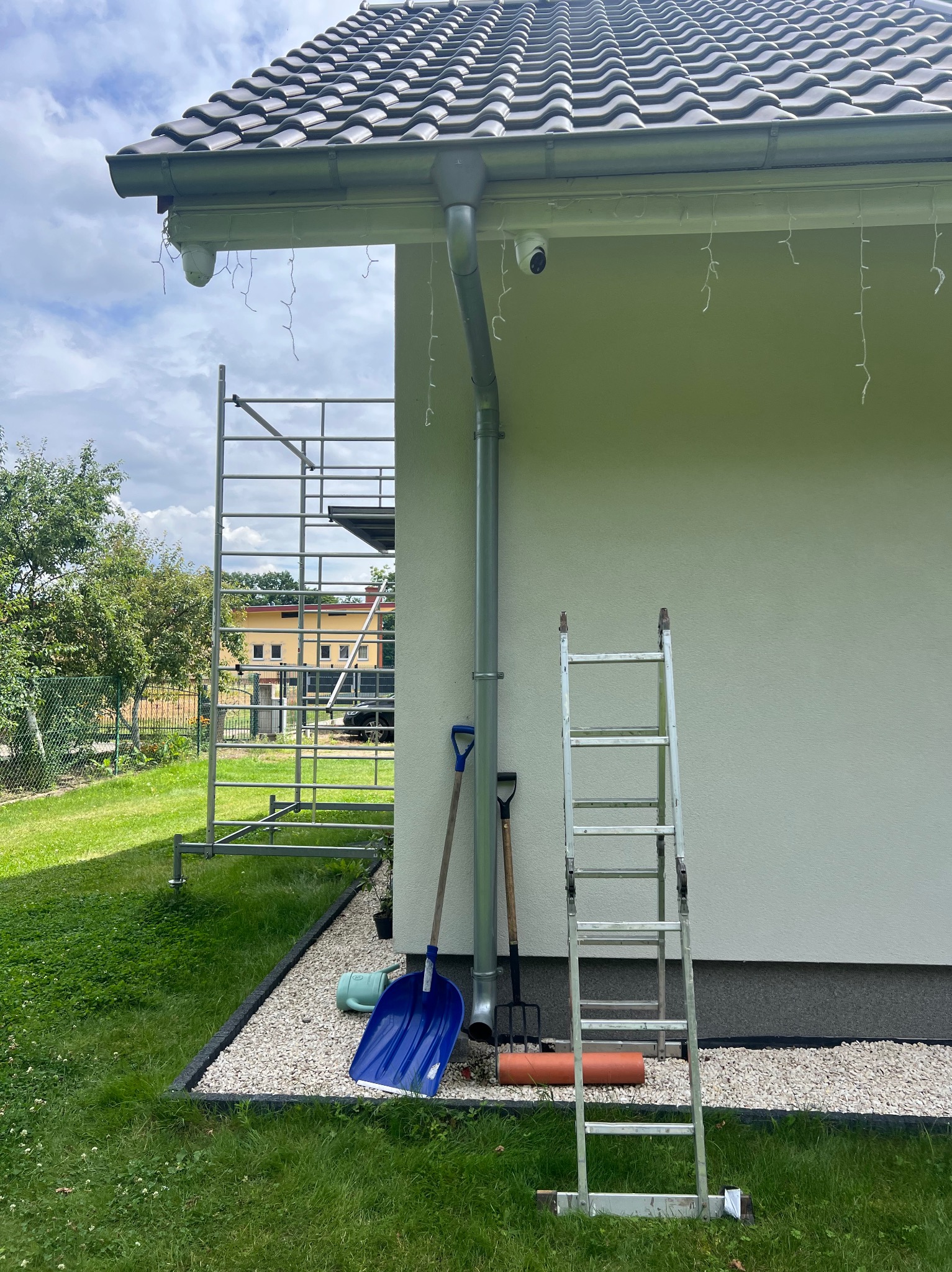 Kamera monitoringu pod dachem domu, obok rynna spustowa. Na trawie drabina, szpadel i widełki. Widoczne rusztowanie i fragment elewacji.