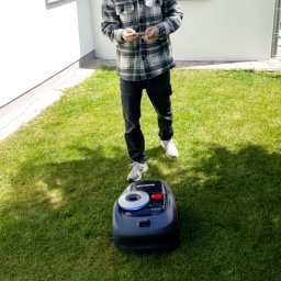 H2OGRODY - Mężczyzna w kraciastej koszuli obsługuje smartfonem automatyczną kosiarkę Navimow na trawniku przed białym budynkiem. Zielona trawa, nowoczesny sprzęt.