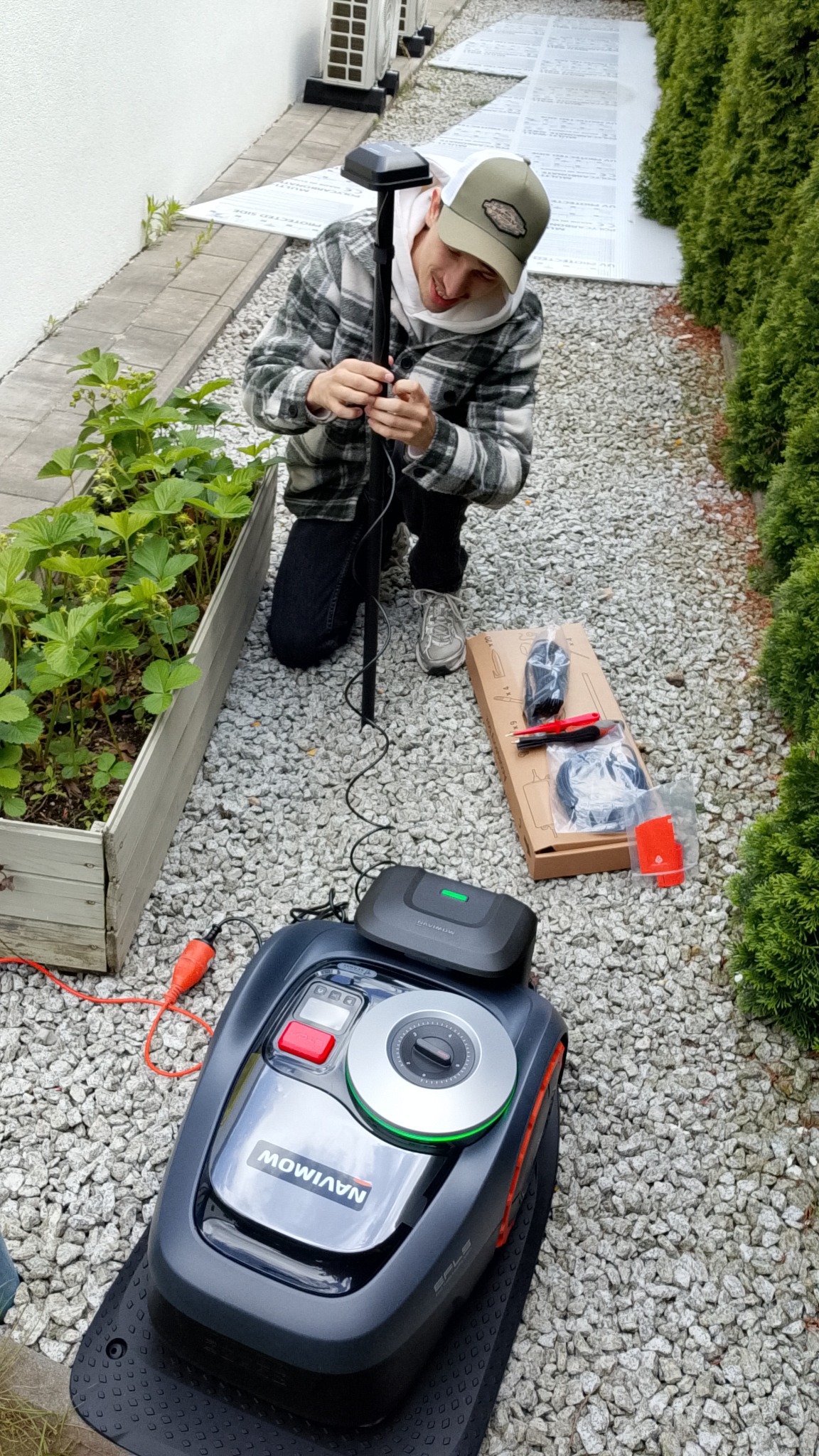 Instalacja robota koszącego Navimow w Lesznie. Mężczyzna montuje stację bazową obok rabaty. Widoczny robot, przewody i akcesoria montażowe na żwirowym podłożu.