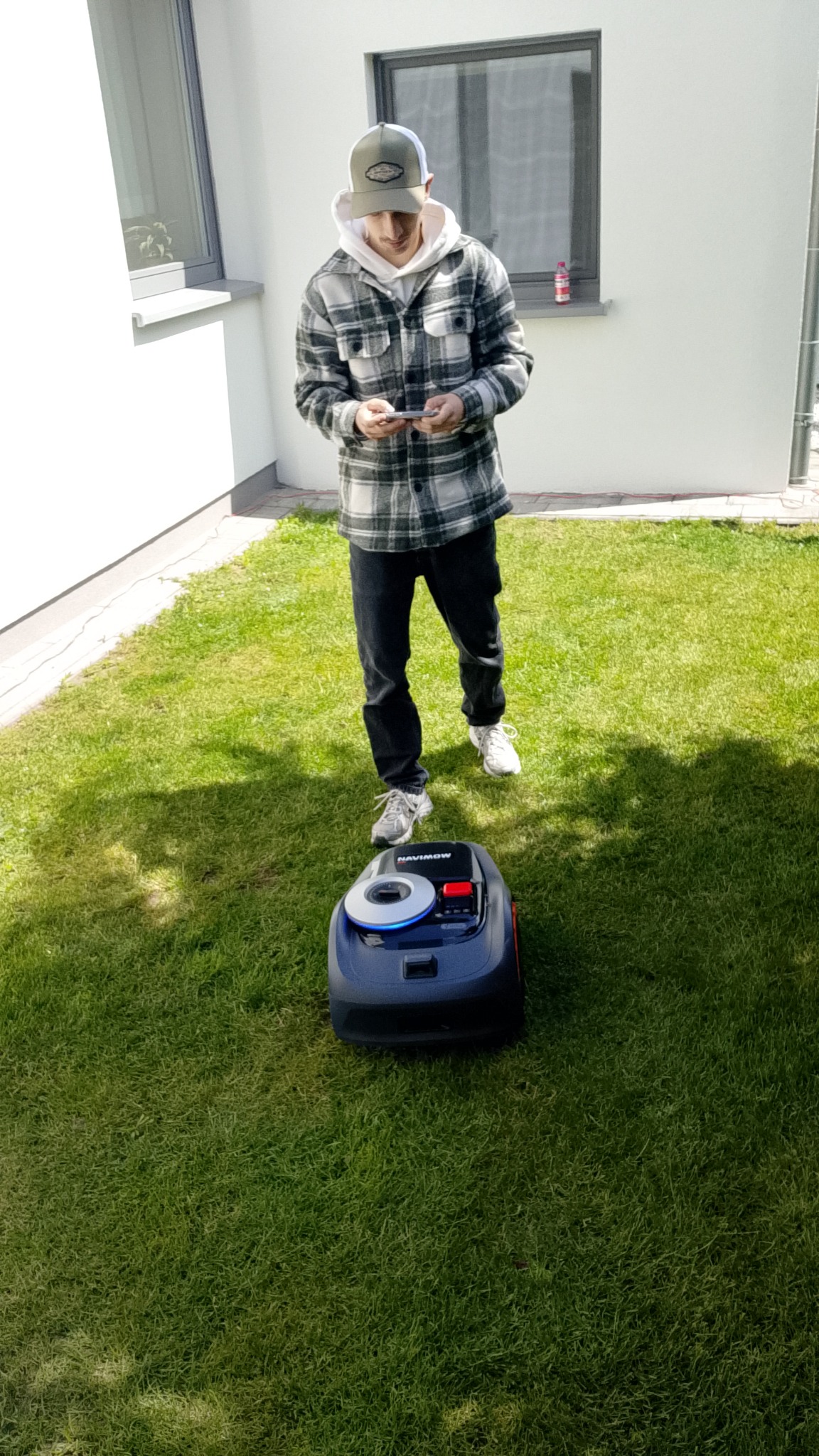 Mężczyzna w kraciastej koszuli obsługuje smartfonem automatyczną kosiarkę Navimow na trawniku przed białym budynkiem. Zielona trawa, nowoczesny sprzęt.
