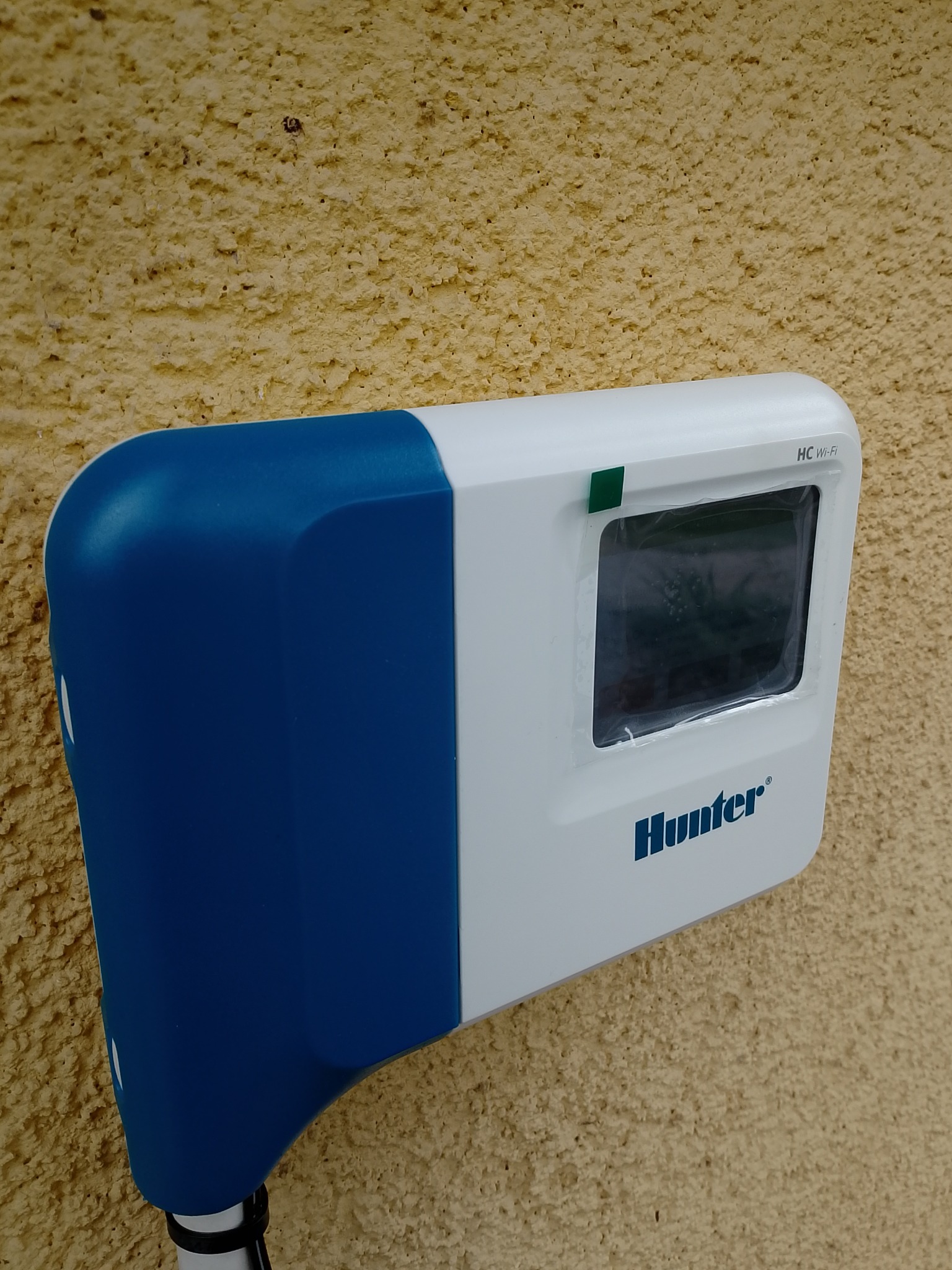 Zamontowany na chropowatej ścianie sterownik Hunter HC Wi-Fi w obudowie biało-niebieskiej. Widoczny wyświetlacz i logo producenta.
