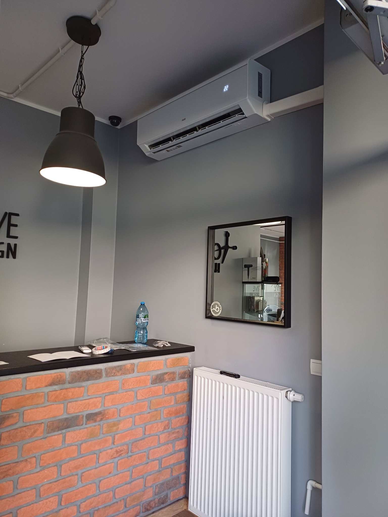Biała klimatyzacja na szarej ścianie nad biurkiem z ceglanym frontem i lustrem w czarnej ramie. Widoczny fragment lampy i grzejnik.