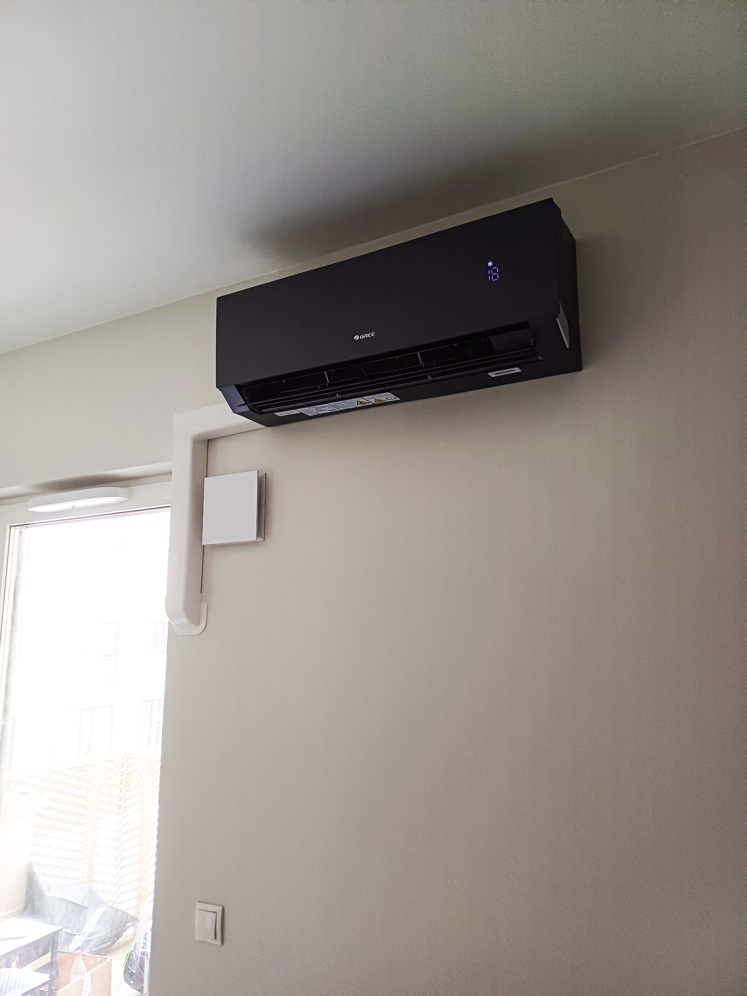 Nowoczesny, czarny klimatyzator Gree zamontowany na jasnej ścianie w pokoju, widoczne białe rury odprowadzające. Widok z dołu, w tle okno.