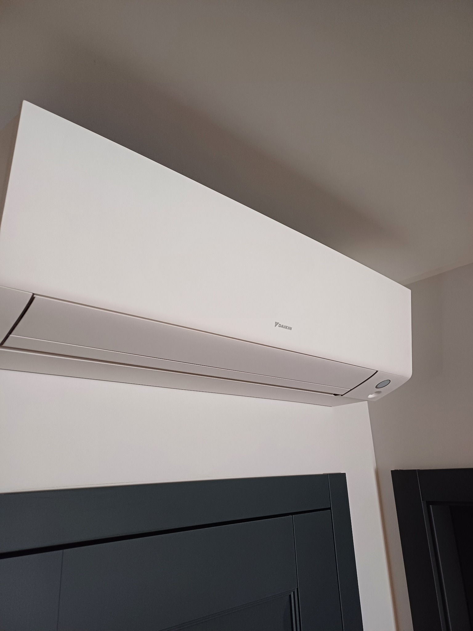 Biała klimatyzacja Daikin na ścianie nad ciemnymi drzwiami, widok z dołu. Nowoczesny design, neutralne tło. Montaż w pomieszczeniu.