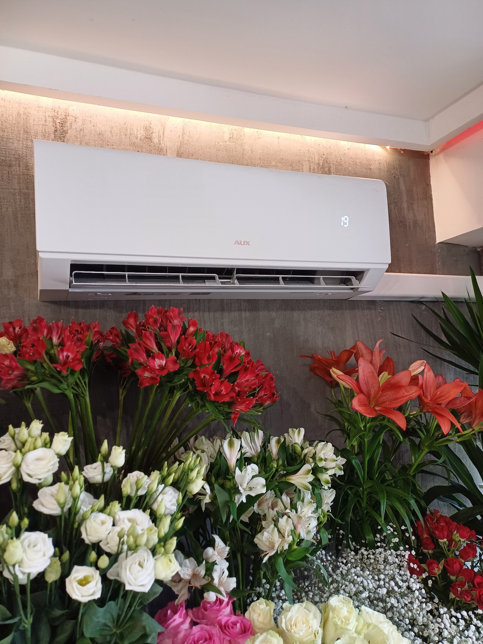 Biała klimatyzacja AUX na ścianie z dekoracyjnym oświetleniem, otoczona bukietami różnorodnych kwiatów w odcieniach czerwieni, bieli i różu.