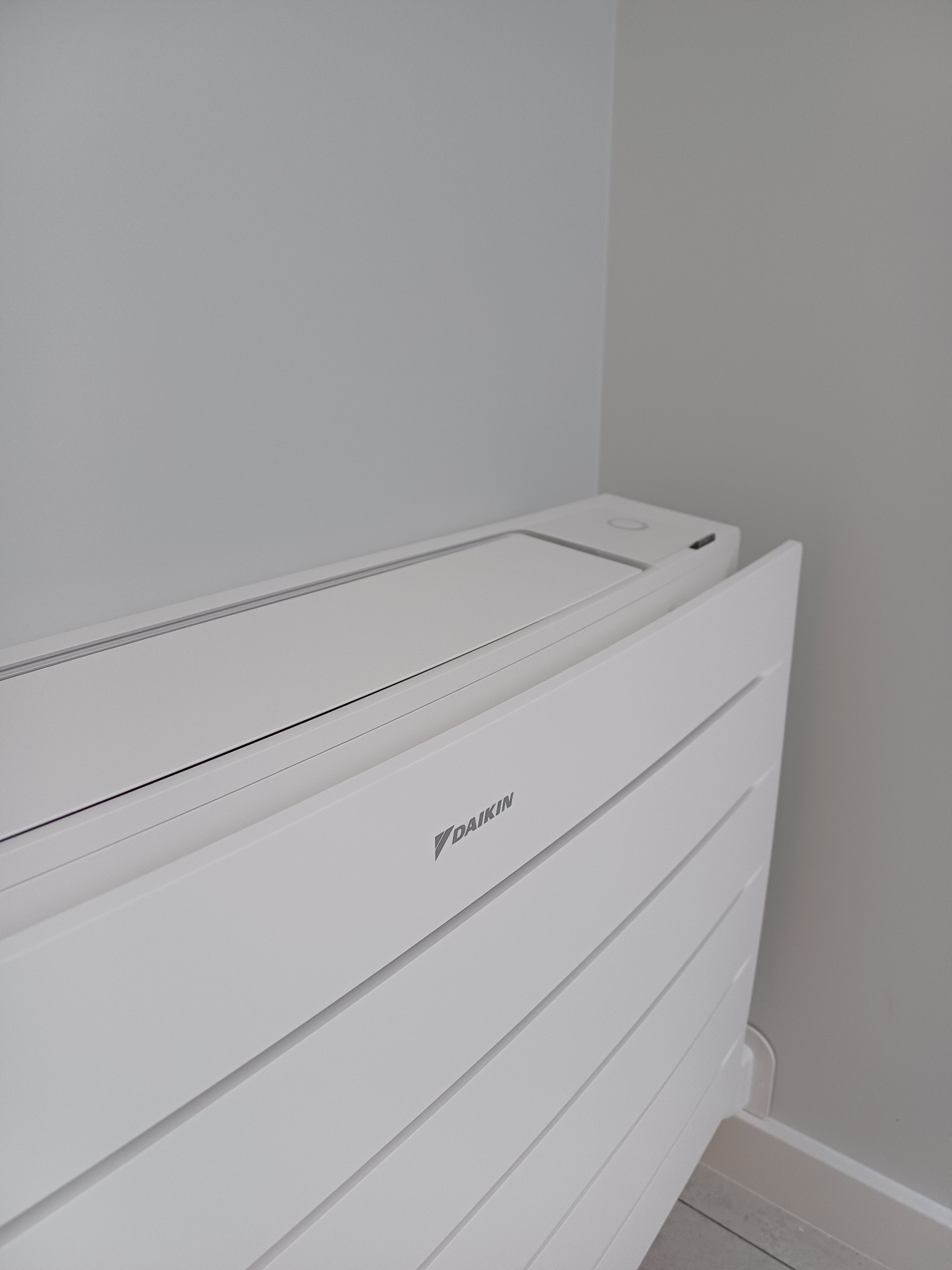 Nowoczesny, biały klimatyzator podłogowy Daikin w rogu jasnego pomieszczenia. Minimalistyczny design z poziomymi panelami i subtelnym logo.